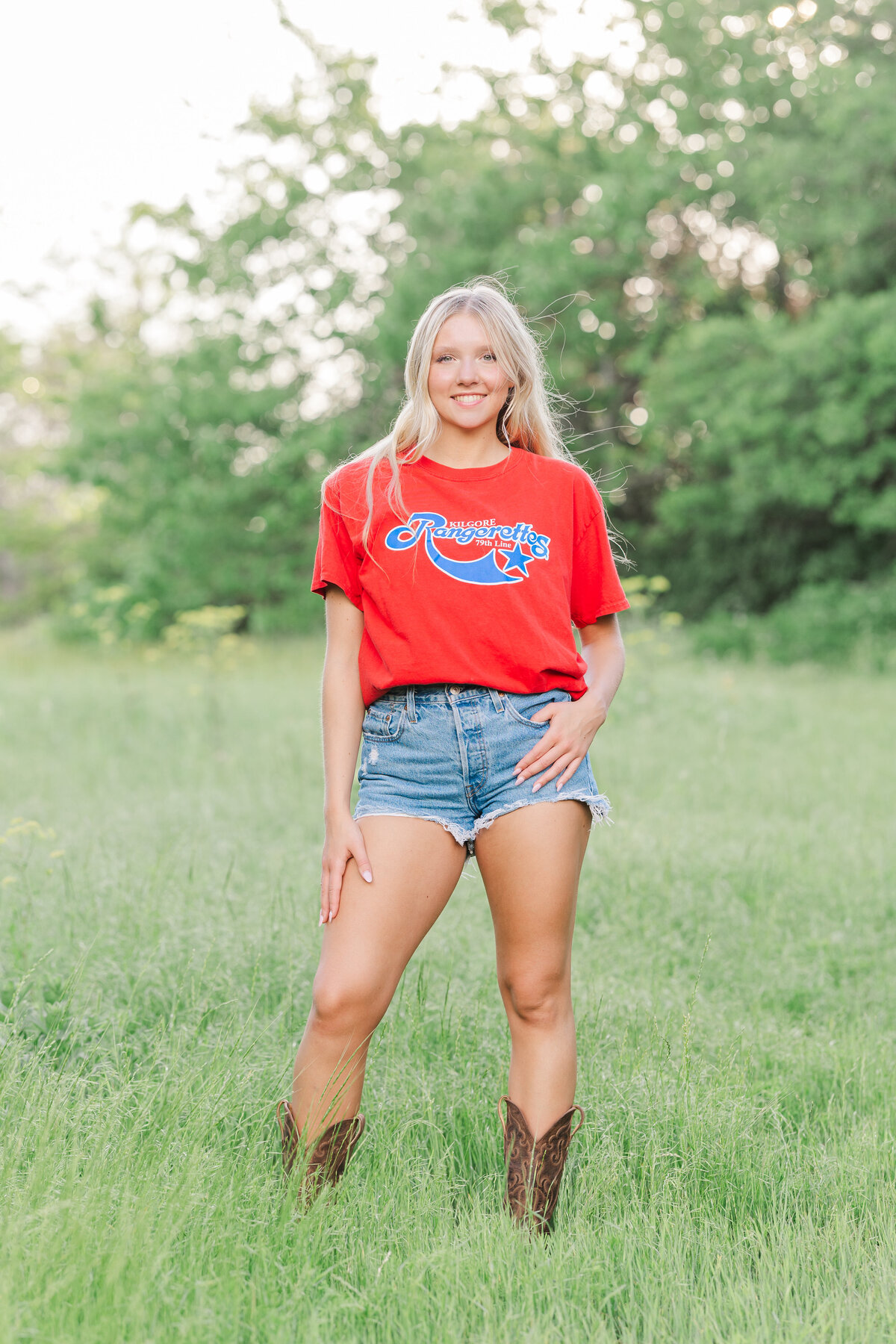 A confident high school senior shows off her casual style in denim shorts, a bold red tee, and cowboy boots in a sunlit field at Arbor Hills in Plano, Texas. Captured by a Dallas area senior portrait photographer.