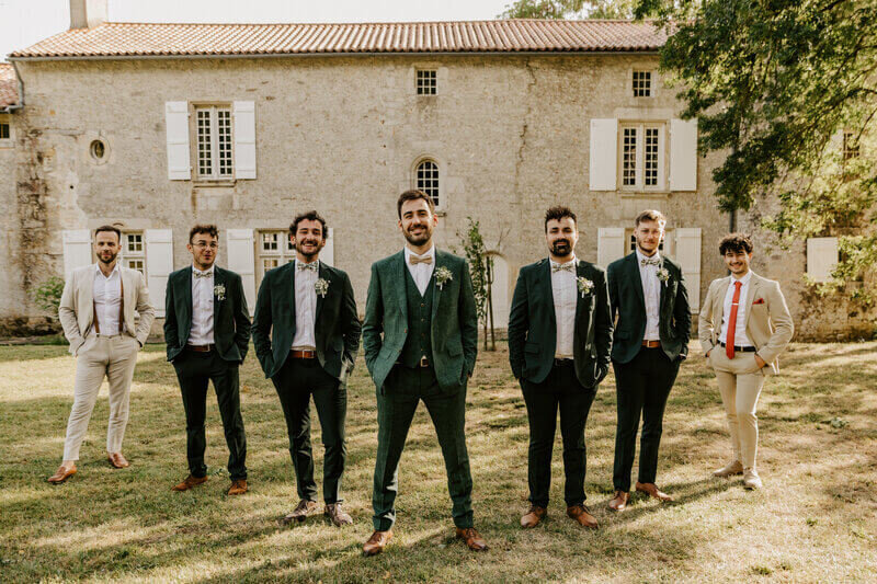 Marié entouré de ses témoins en costume vert et amis en costume beige alignés les uns les autres, prenant la pose devant un bâtiment en pierres pour Laura, photographe mariage en Vendée.