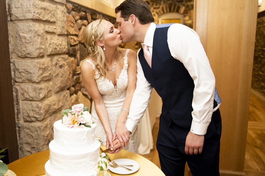 A bride and groom kiss as they pose for a photo while cutting their wedding cake.