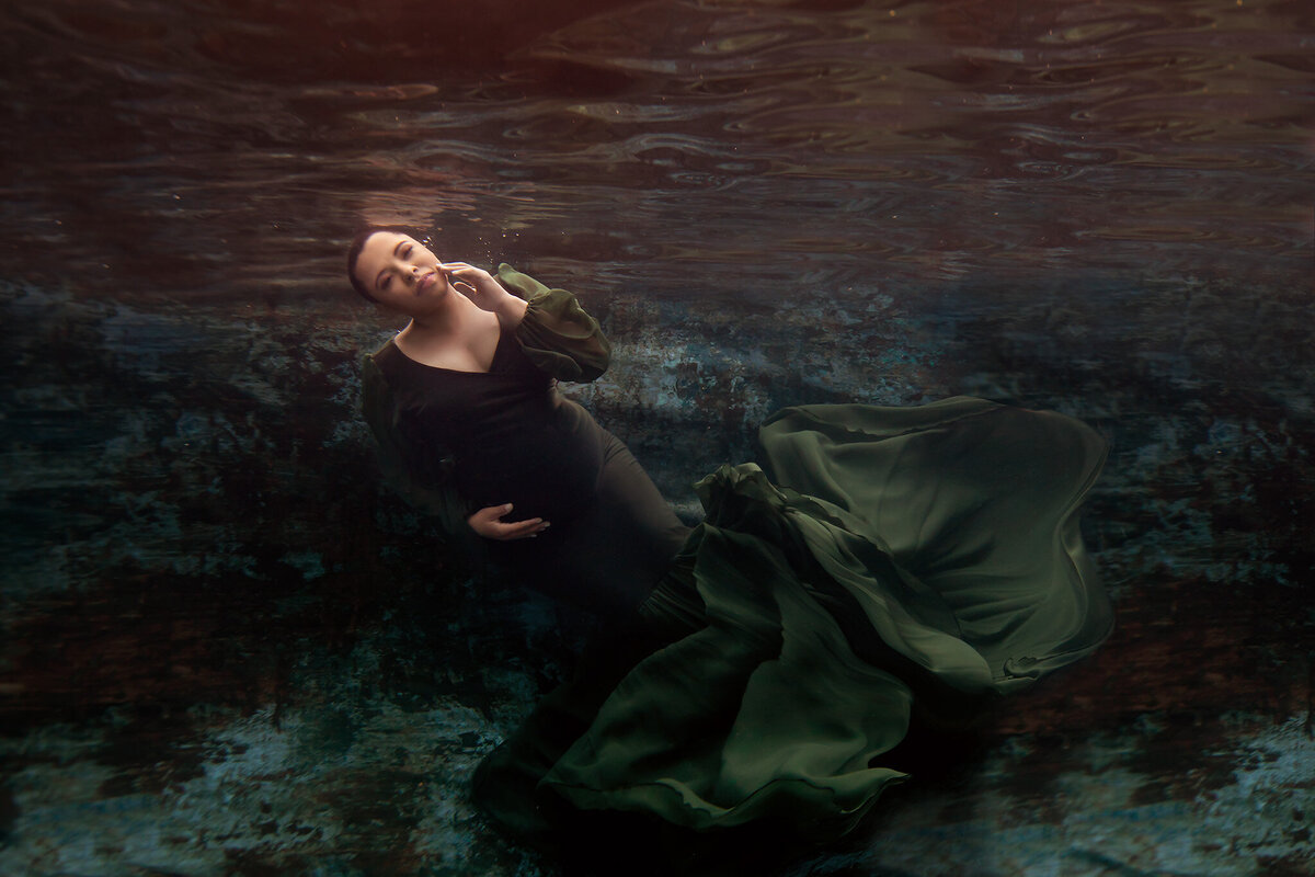 Underwater maternity photo of a woman in a flowing dark green gown resting gracefully against a textured, painterly background. This underwater maternity photoshoot captures her serene expression and the gentle motion of fabric in the water, embodying the artistry of a Dallas underwater maternity photographer.