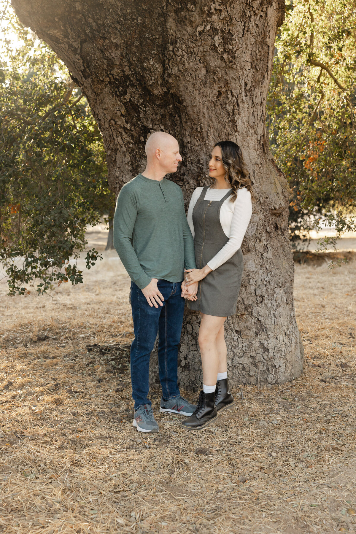 Couple standing together under a tree in rustic outdoor setting, smiling warmly. Photographed by Yucaipa family photographer Kaitlyn Dawn Photography.