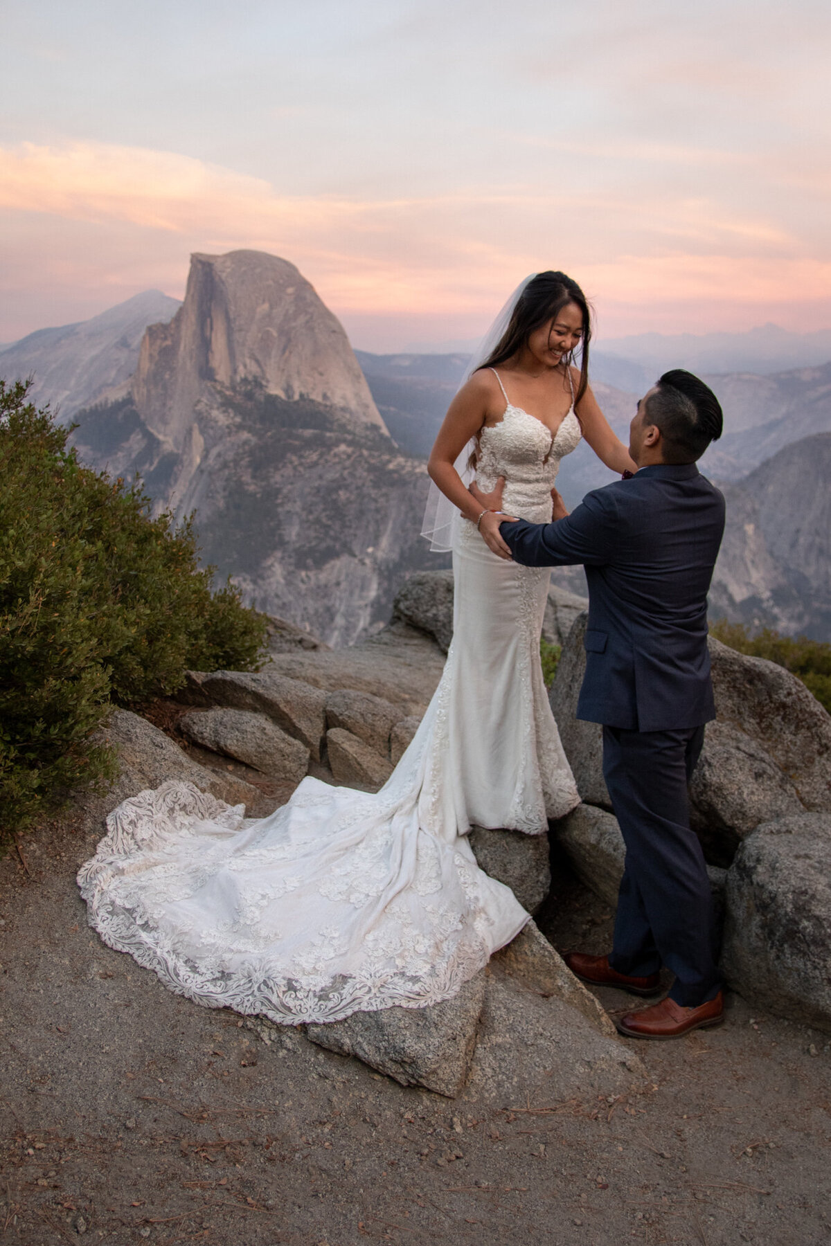 A bride stands on a rock with her hand on her grooms shoulder on their elopement day.