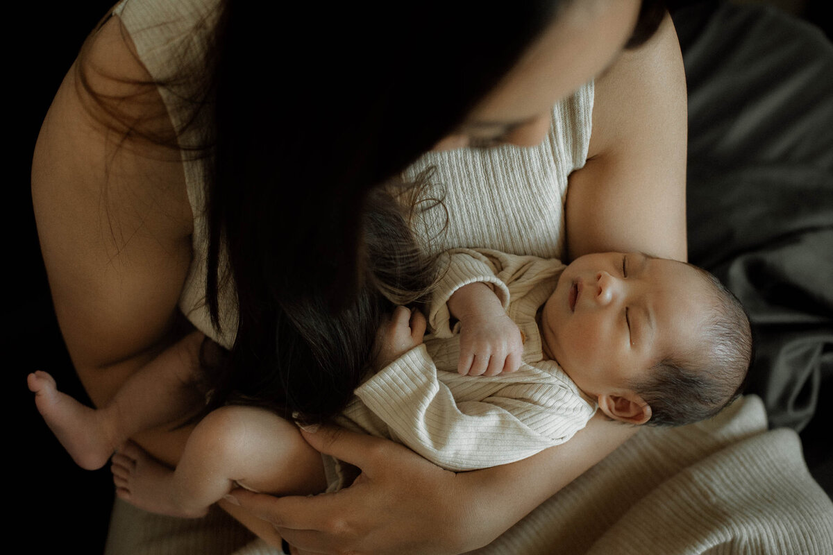 Mother holding her newborn close against her chest, both dressed in neutral tones, as she gazes tenderly at the baby resting in her arms.