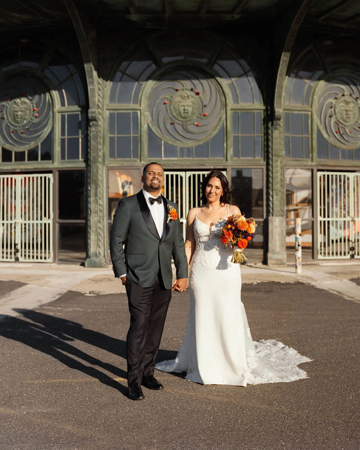A bride and groom stand hand-in-hand outdoors, smiling, captured beautifully by a film photographer NJ. The groom wears a dark green tuxedo, the bride holds vibrant flowers, and an ornate building with arched doors stands behind them.
