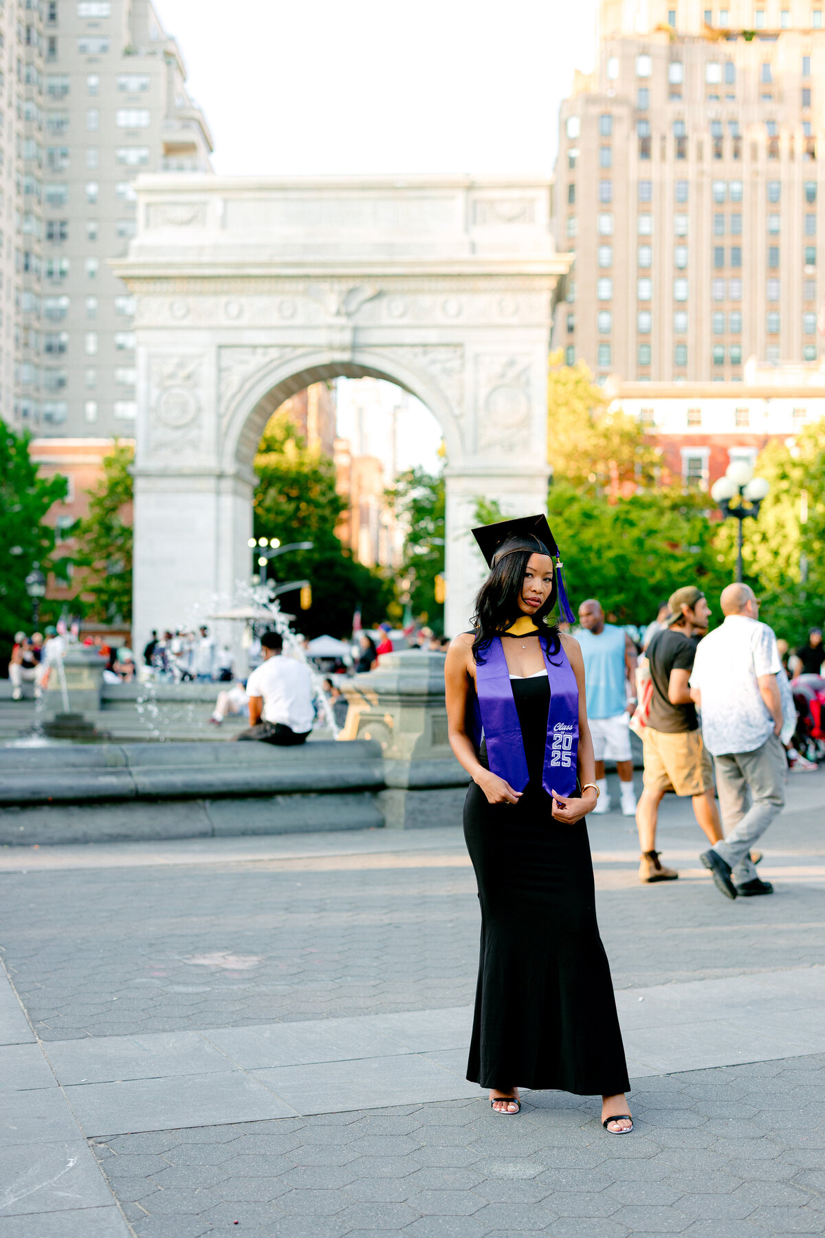 NYU graduate smiling in black dress and purple stole at Washington Square Park.