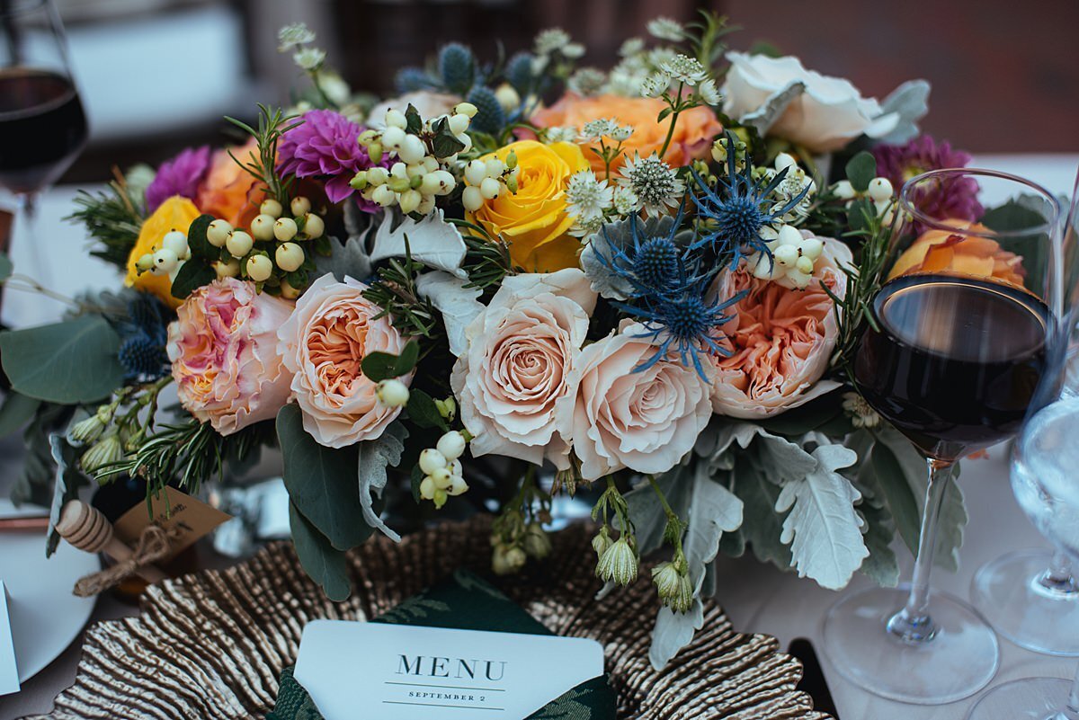 Large floral centerpiece with blush, peach, yellow, purple and pink roses, blue thistle, dusty miller and seeded eucalyptus on a reception table for a wedding at Cheekwood