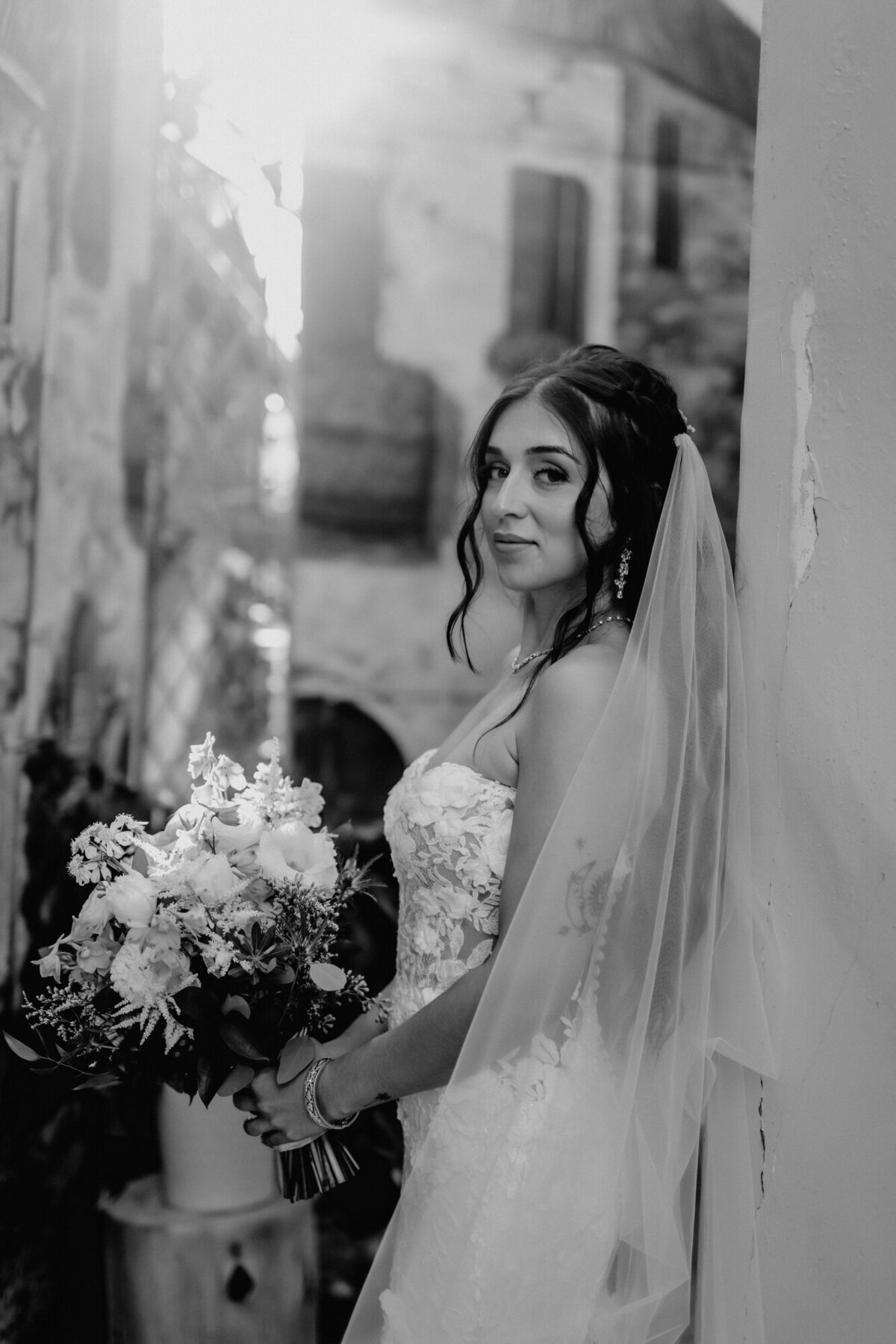 Black and white photo of the bride leaning agaist the wall holding her bouquet and looking at the camera over her shoulder.