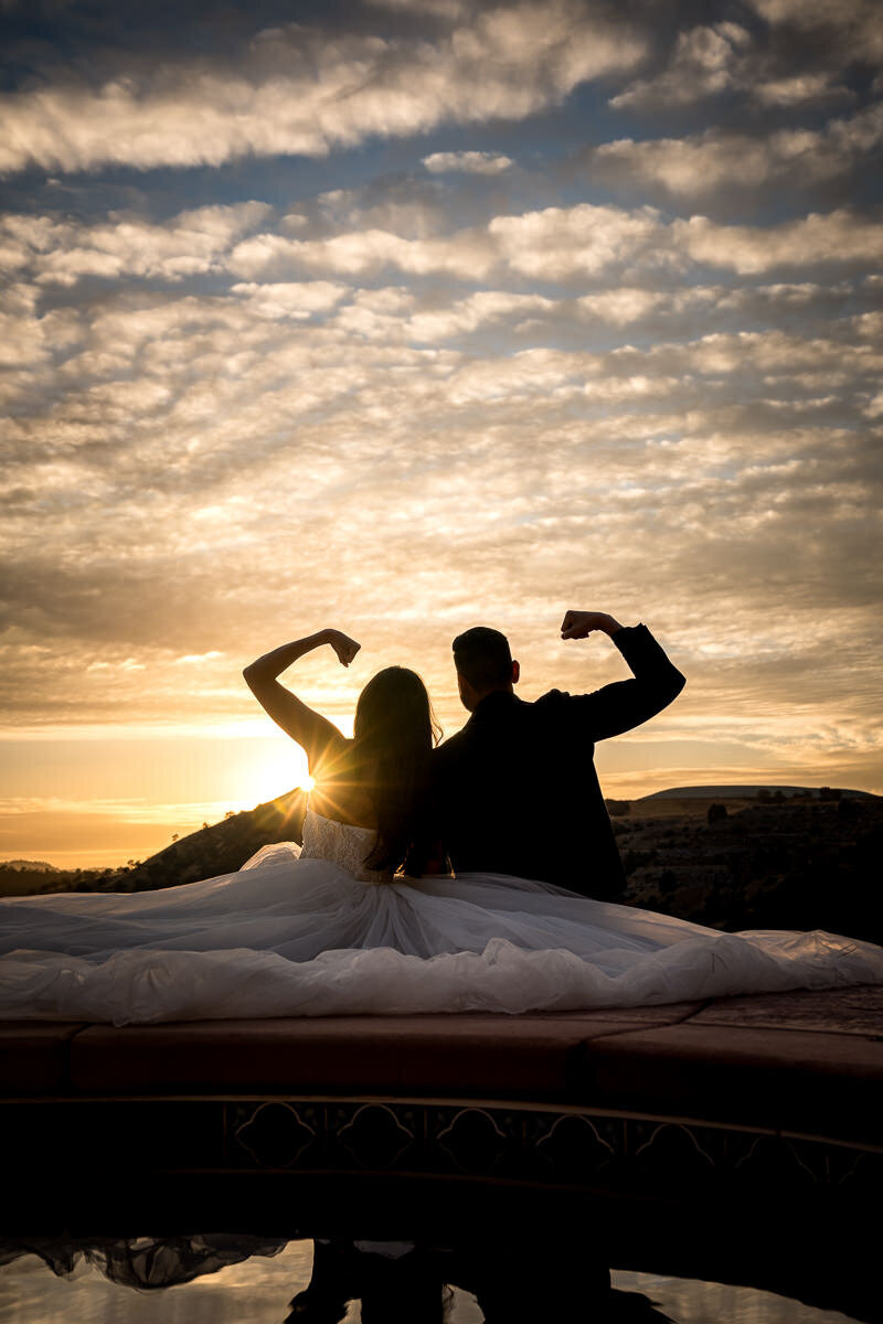 A fun and lighthearted wedding moment as the bride and groom flex their muscles while admiring a spectacular sunset at Airola Road Vineyard, celebrating their love with laughter and stunning views.