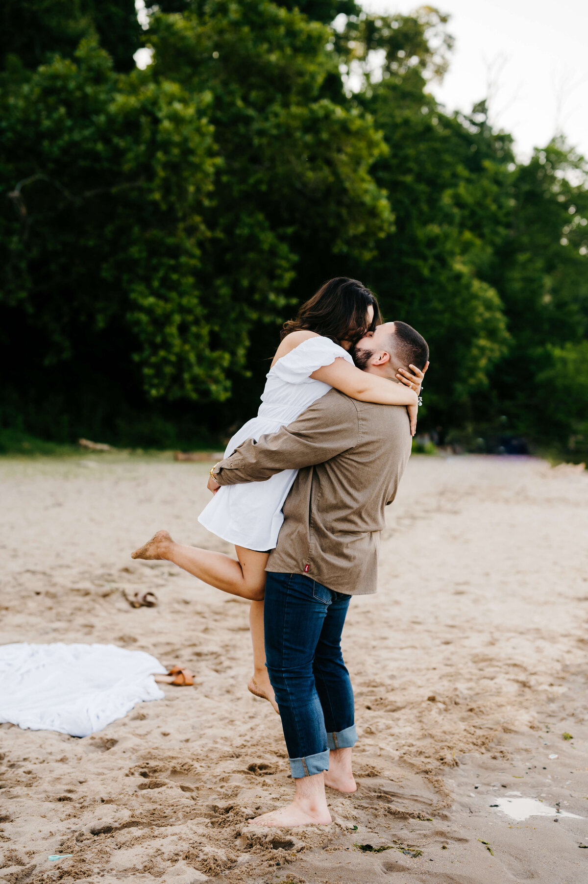 white dress for engagement photos on a dark haired woman as her fiance holds her on the beach with a tree line in the distance