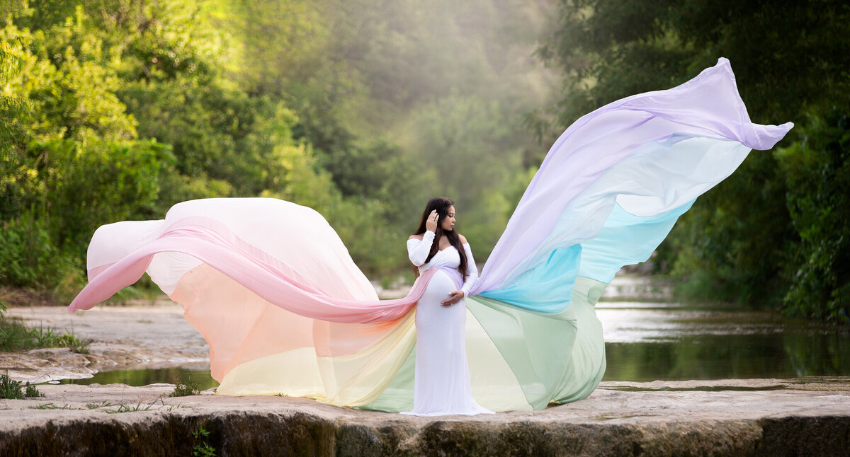 Dramatic maternity image with flowing rainbow fabric dress by a creek in Cedar Park, TX