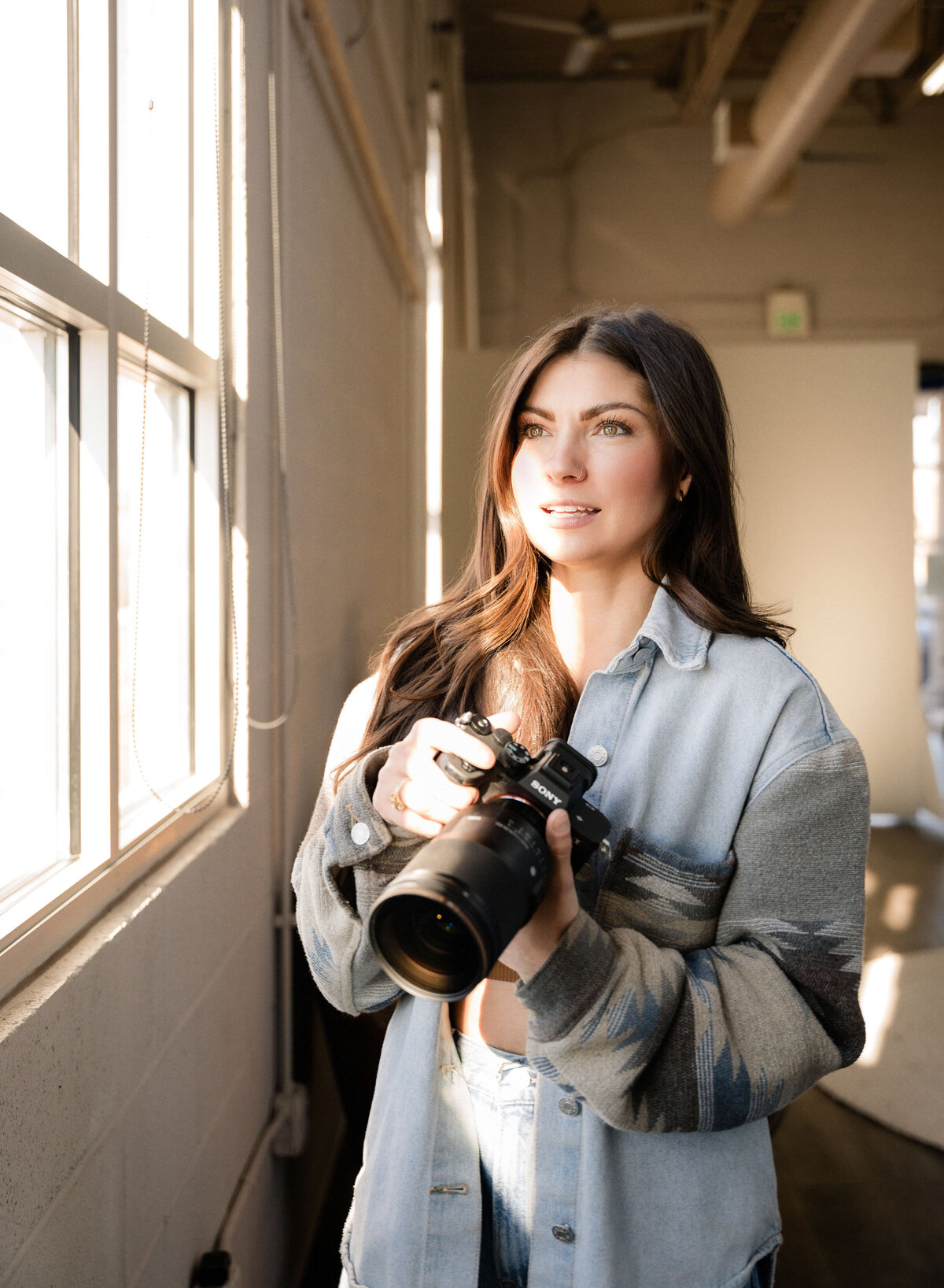 A young Photographer stands, smiling, while looking out a window where the light spills in on her face