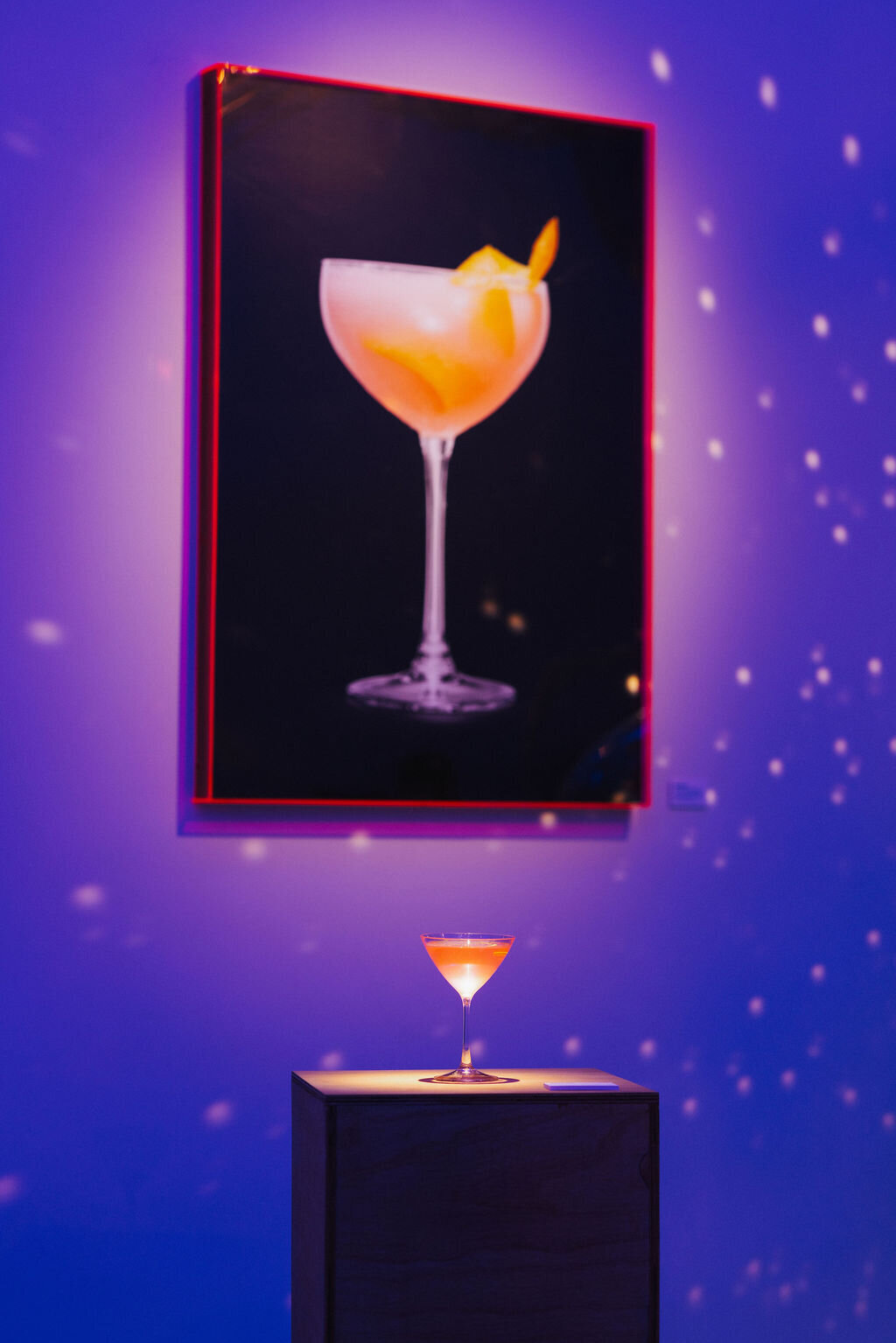 Cocktail on display under matching framed artwork in a glowing blue Absolut Vodka × Like Minded Creatures installation