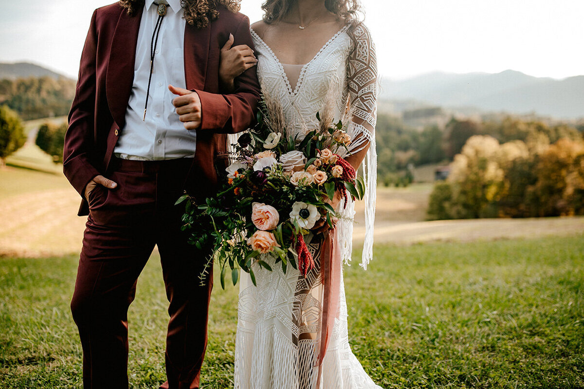 Bride and groom pose side by side locked in arms wearing a white wedding gown and burgundy tuxedo with bride holding bouquet