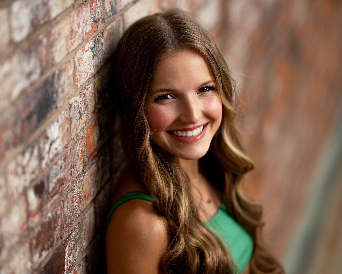 A brunette in a green dress leans on a brick wall smiling