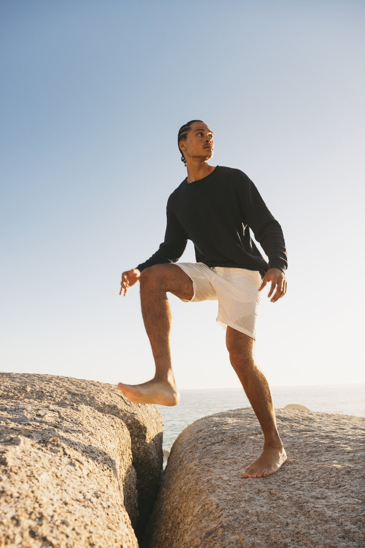 Male model Timothe stepping confidently between two large rocks at the coast in Cape Town, dressed in a black sweater and white shorts, under clear blue sky.
