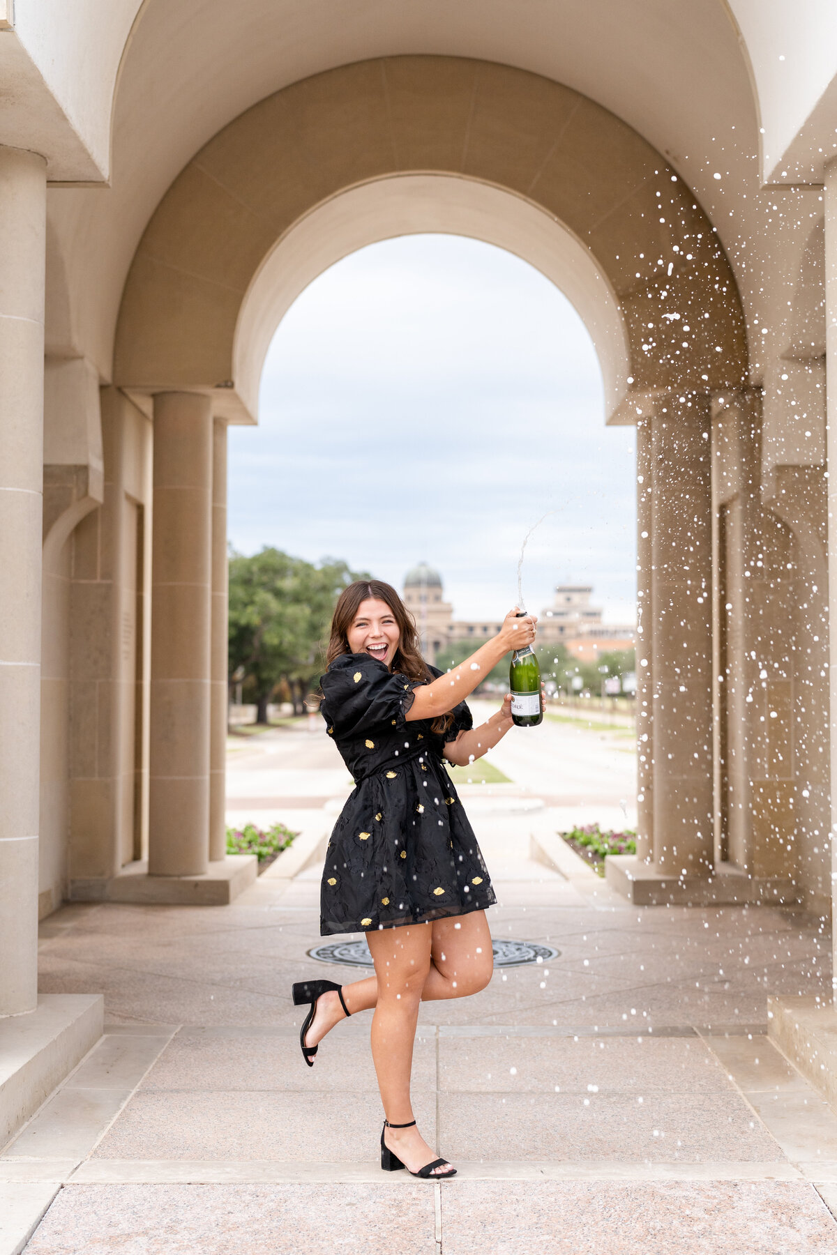 Texas A&M senior girl wearing short black dress and heels spraying champagne and celebrating in the archways of the Bell Tower
