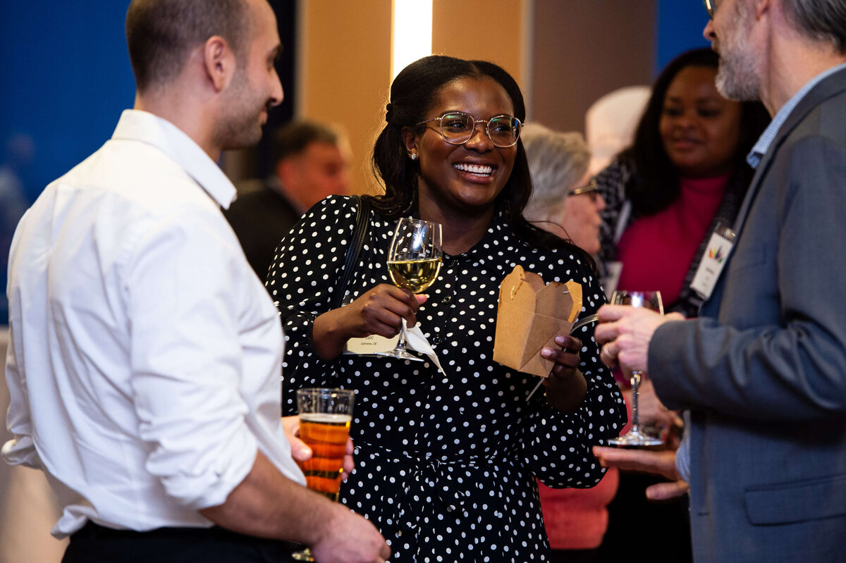 a black woman holding a glass of wine and smiling with attendees during a corporate reception.  Captured by Ottawa Event Photographer JEMMAN Photography COMMERCIAL