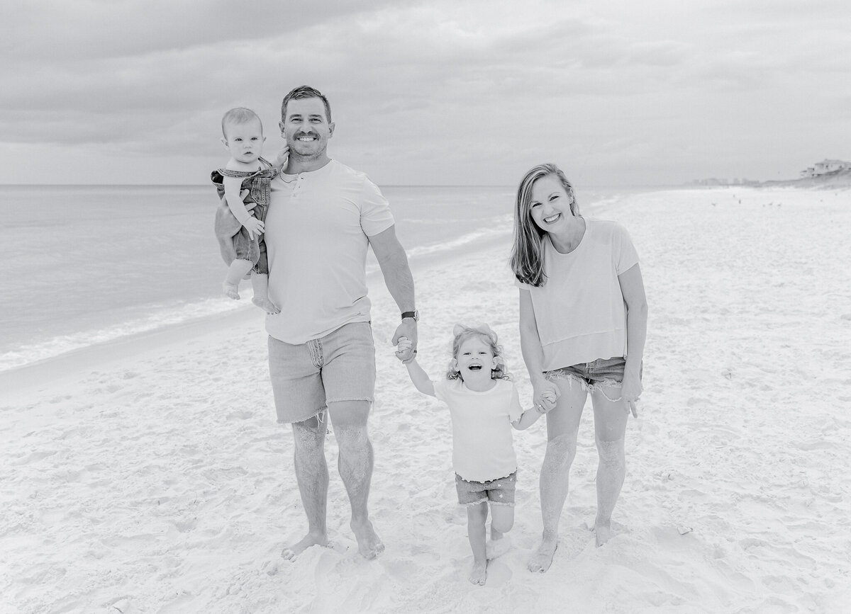 A happy family of four poses on the white sandy shores of Orange Beach, AL. The father, dressed in a neutral polo and denim shorts, carries his young daughter on his shoulders as she grins with a pink bow in her curly hair. The mother, smiling in a beige top and denim shorts, cradles a baby in denim overalls. The turquoise waves and soft clouds create a stunning backdrop for this cherished beach moment.