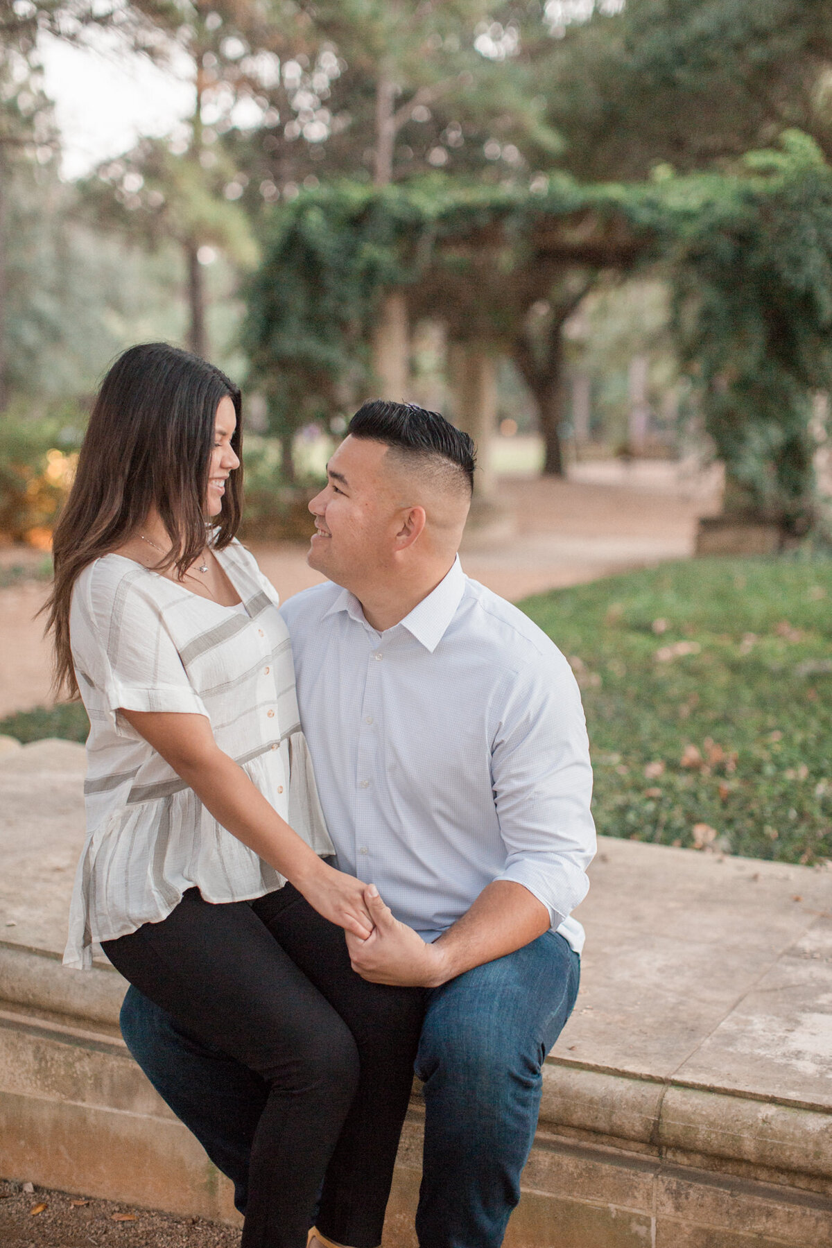 A engaged latino couple beam at each other while she sits on his knee in Hermann Park