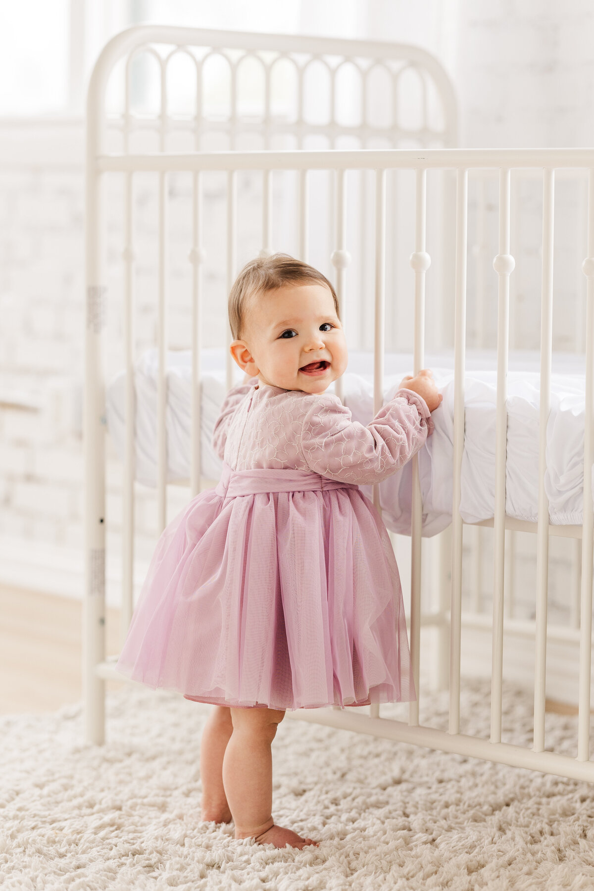 baby-girl-wearing-a-blush-velvet-dress-standing-by-crib-at-the-lumen-room-studio