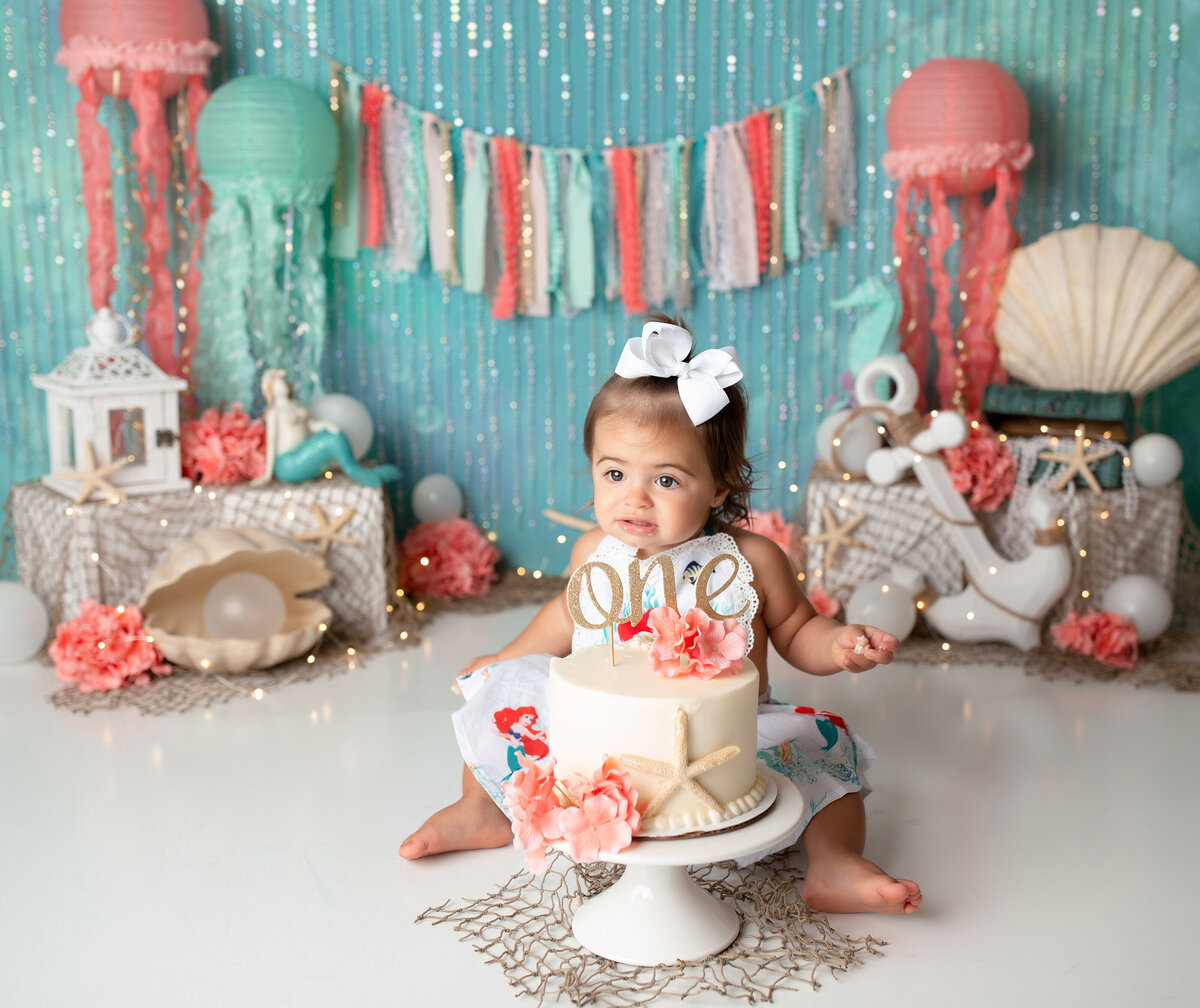 Under the Sea themed cake smash in West Palm Beach and Delray Beach photography studio. Baby girl is sitting behind a white cake decorated with seashells and coral flowers. She is looking off in the distance. In the background, there are jellyfish, mermaid, and under the sea decor.