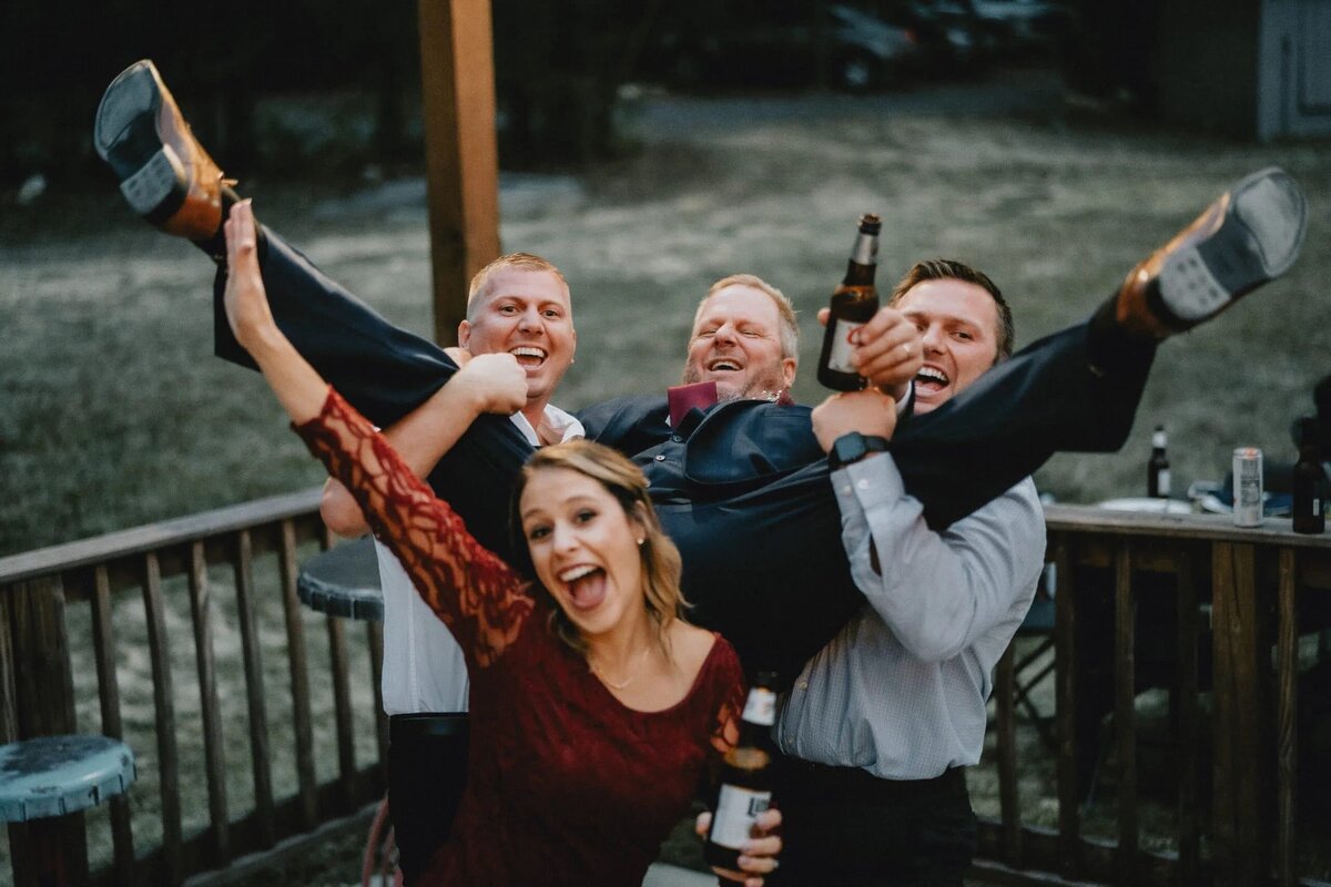 A group of wedding guests and groomsmen lift the groom into the air, cheering with drinks in hand