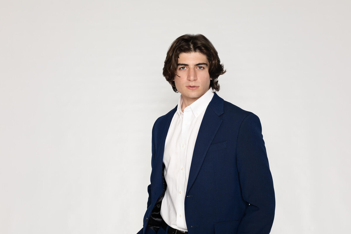A person with medium-length brown hair wearing a navy blue blazer and white shirt stands against a plain white background, looking directly at the camera.