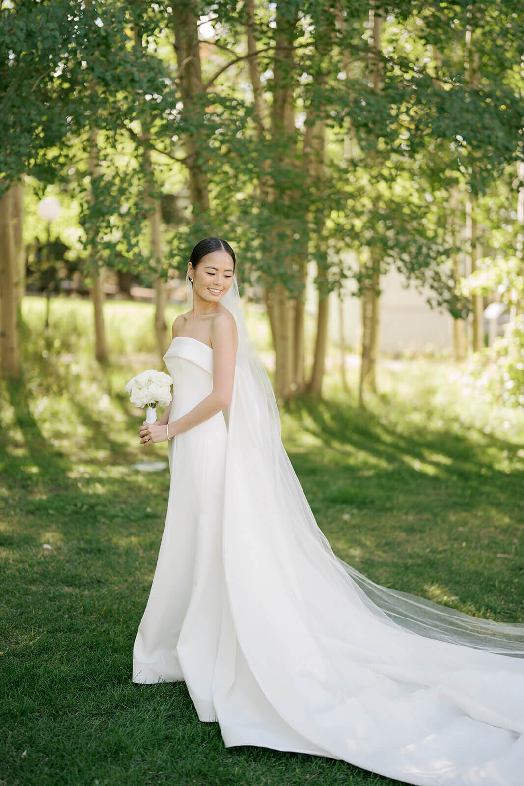 A bride, wearing a simply elegant gown, stands holding her bridal bouquet of white roses among the fairytale Colorado summer greenery at her Aspen Meadows Resort Wedding.