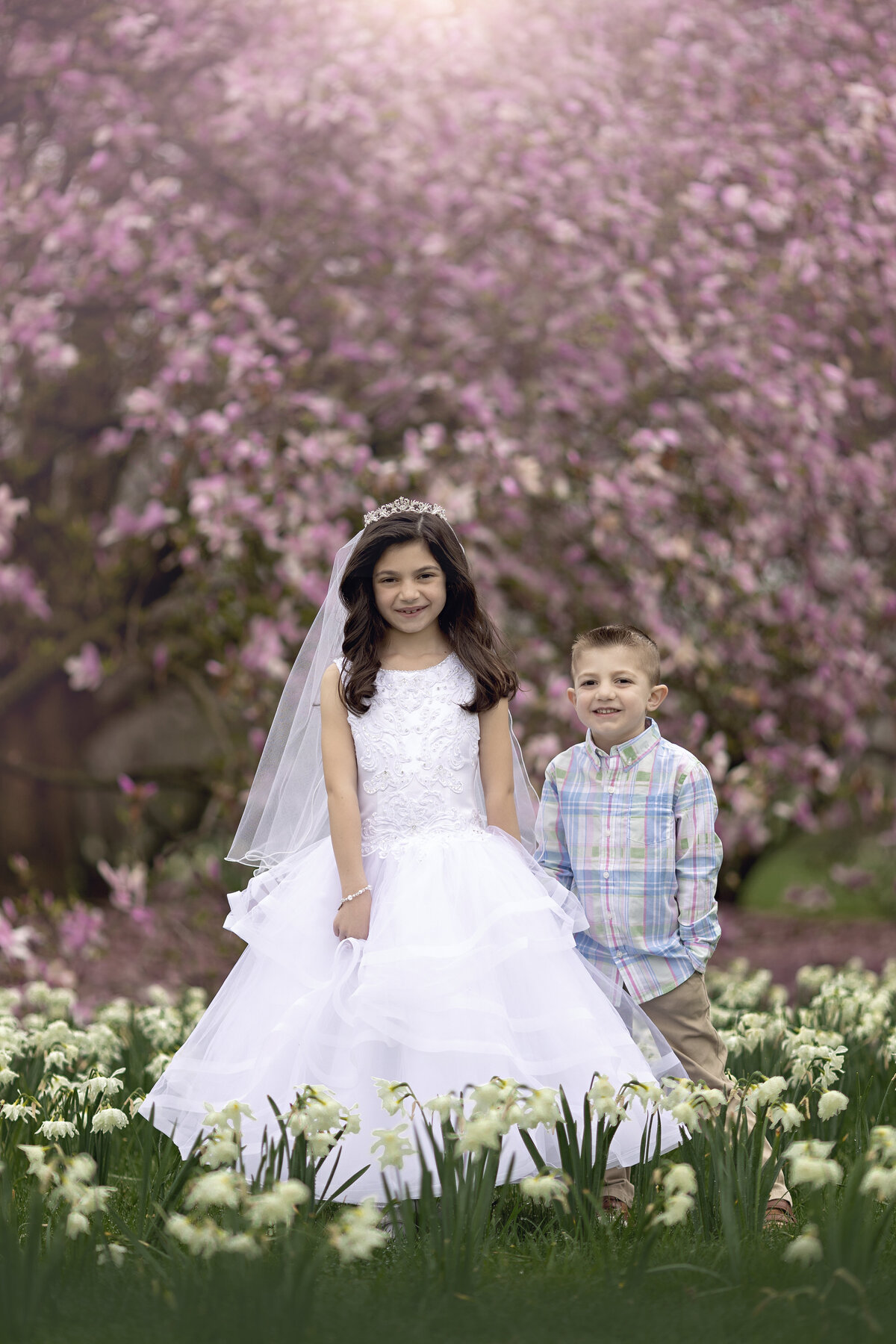 A young girl in a white dress stands with her little brother in a field of daffodils with her toddler brother