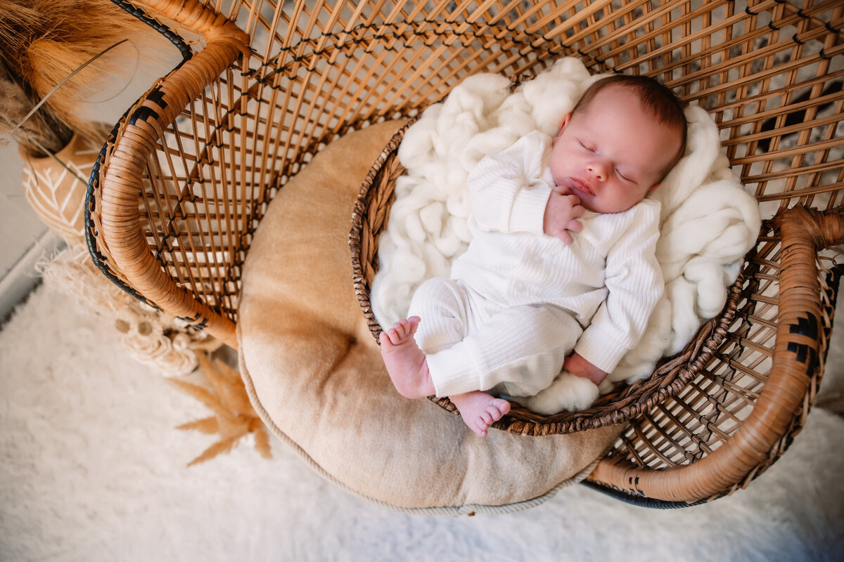 A sleeping newborn baby in a wicker chair wearing a white onesie