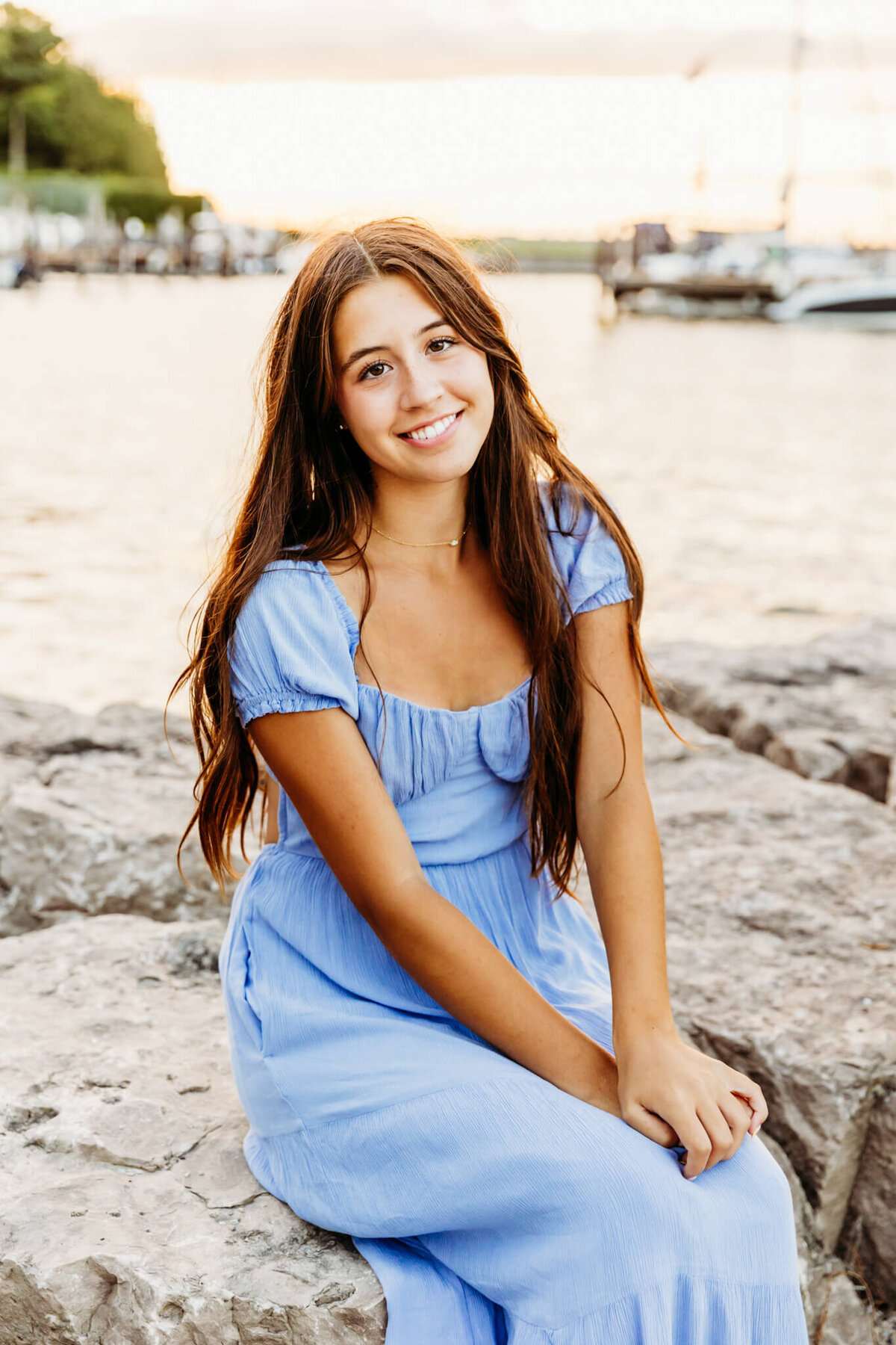 gorgeous brunette senior in a blue dress sitting on large rocks alongside the lake at sunset captured by Ashley Kalbus Photography