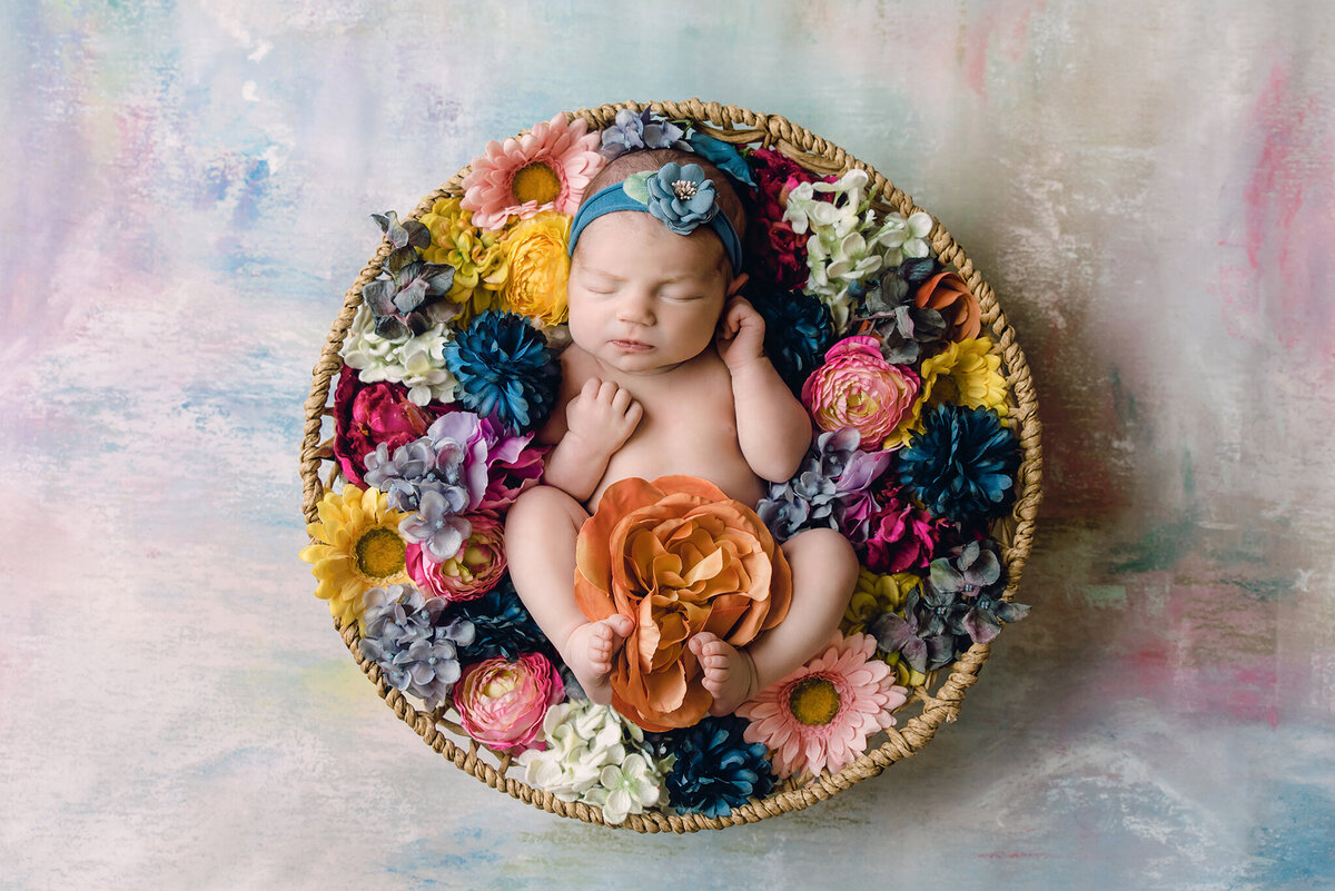 baby girl sleeping in round basket surrounded by different color flowers