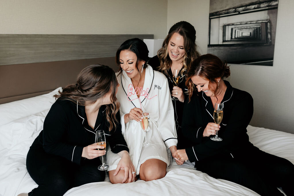 Bridal party getting ready at the Palm Event Center in Pleasanton, CA. The spring wedding scene shows the bride in white pajamas and bridesmaids in black pajamas, all sitting on a bed and laughing.