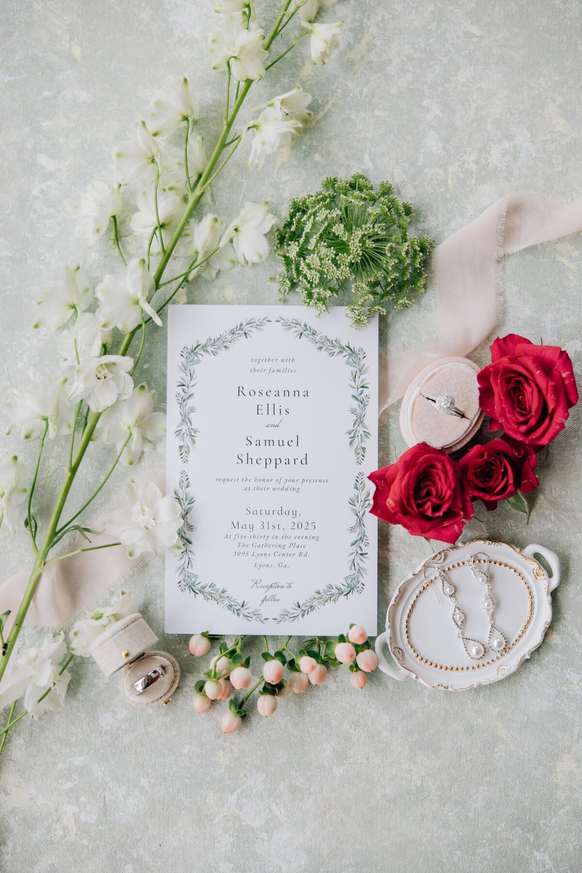 flat lay of a wedding invitation surrounded by greenery, red roses, and other wedding details