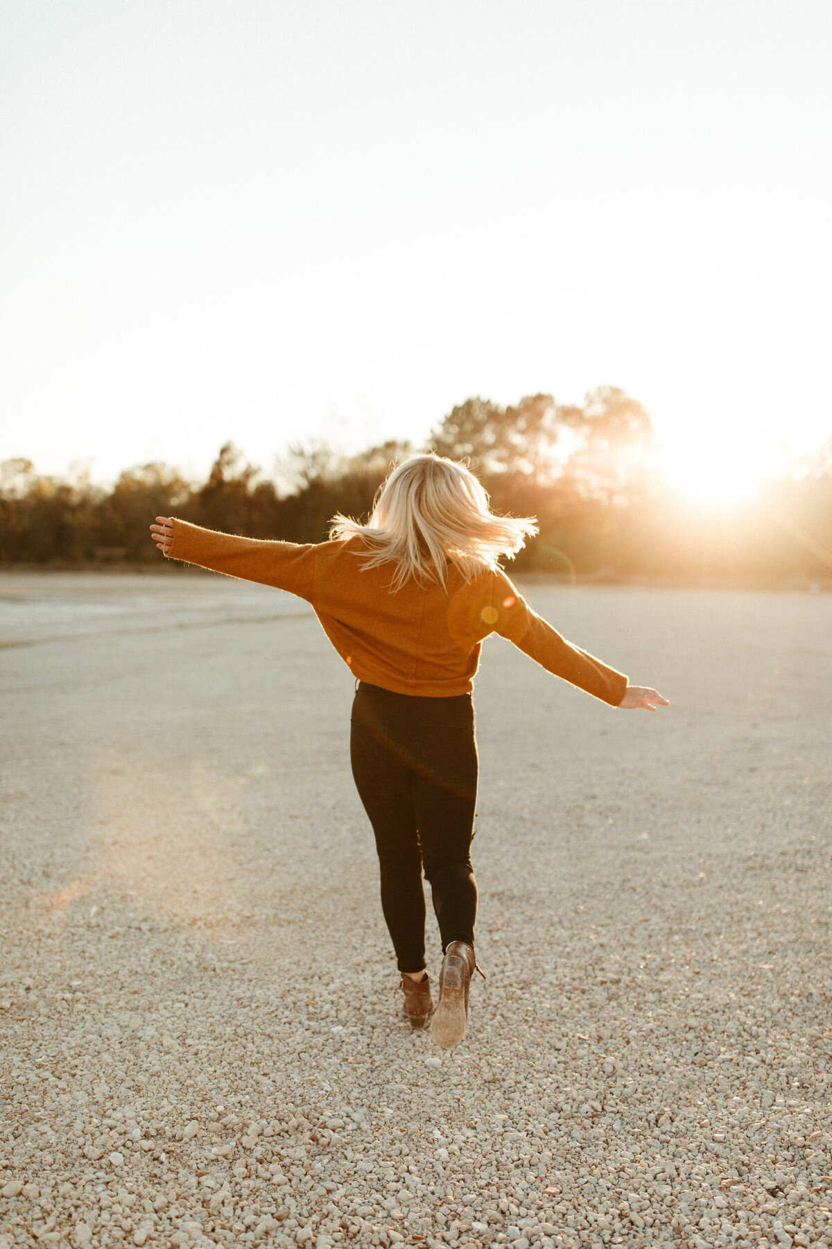 High school senior in baggy sweater running and twirling in a big open gravel area with the sunset behind her