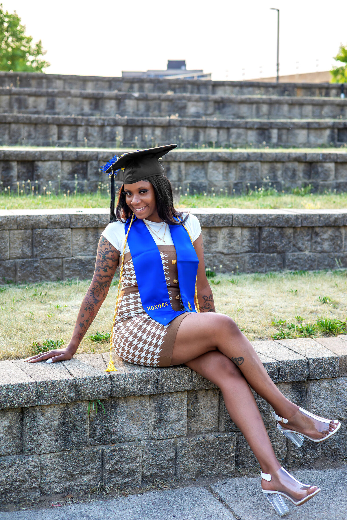 College graduate wearing graduation cap, blue honors sash, yellow cord and brown and white dress sitting on a concrete ledge
