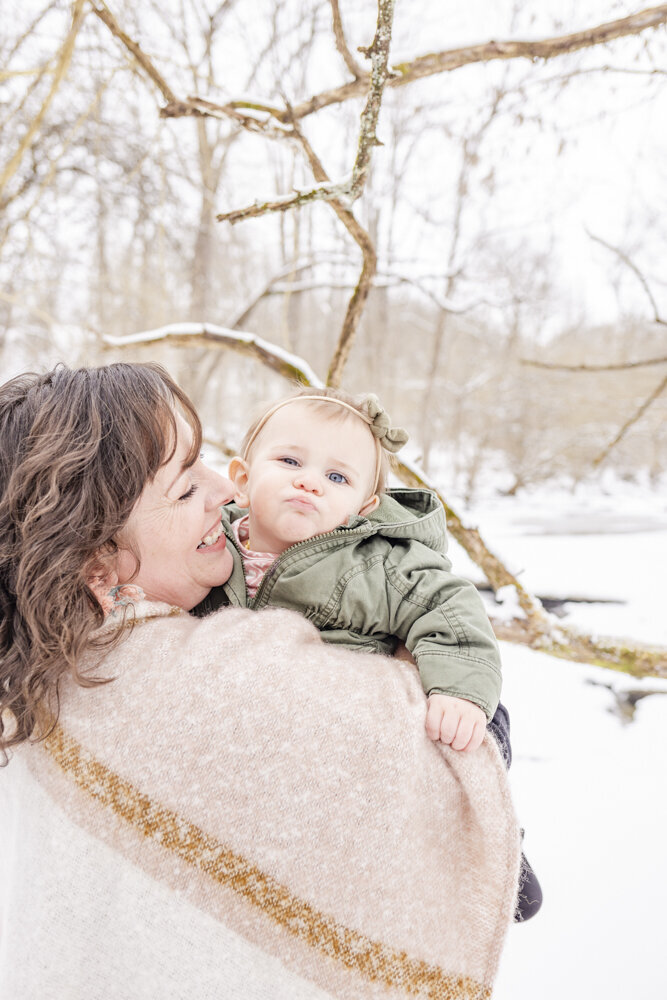 Mom smiling with baby daughter who is making a silly face in Russellville, Ohio on a cold, snowy, Winter day.