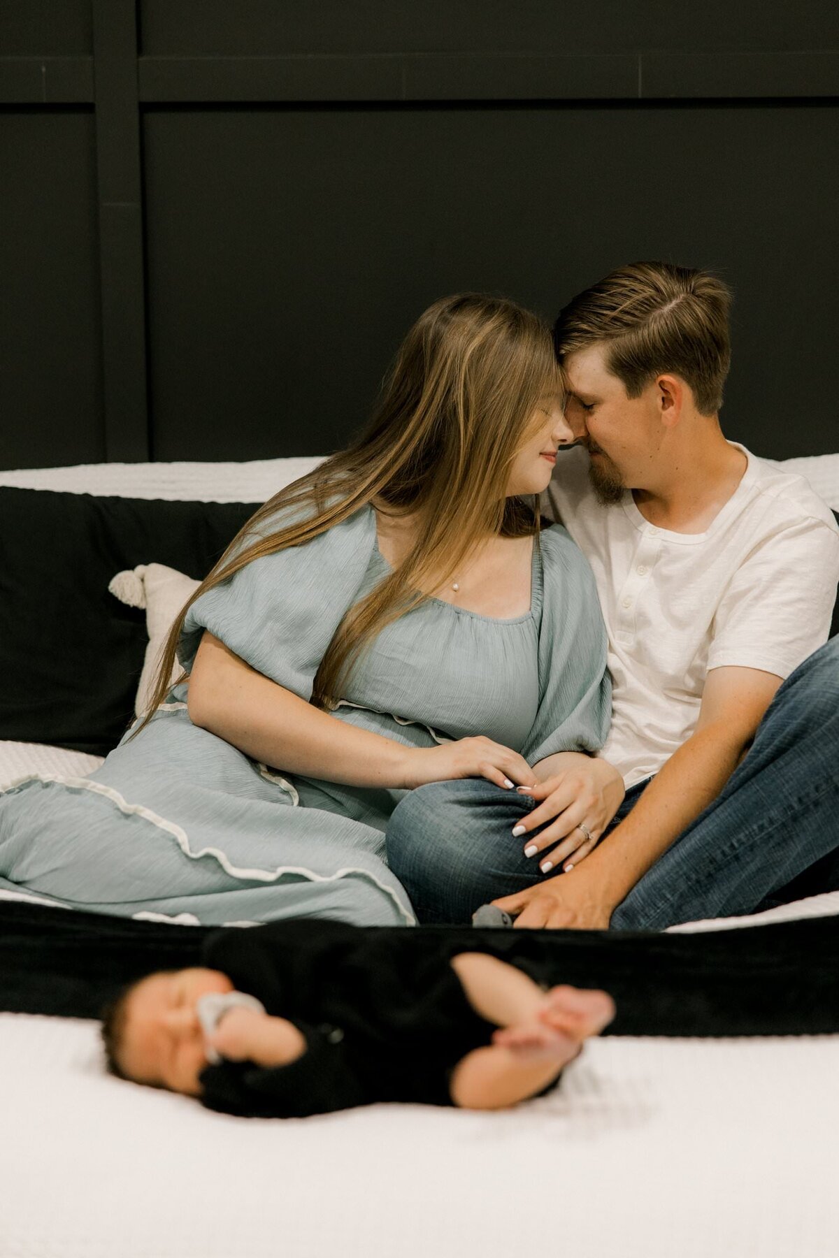 couple sitting in bed foreheads touching baby at the front of the bed 