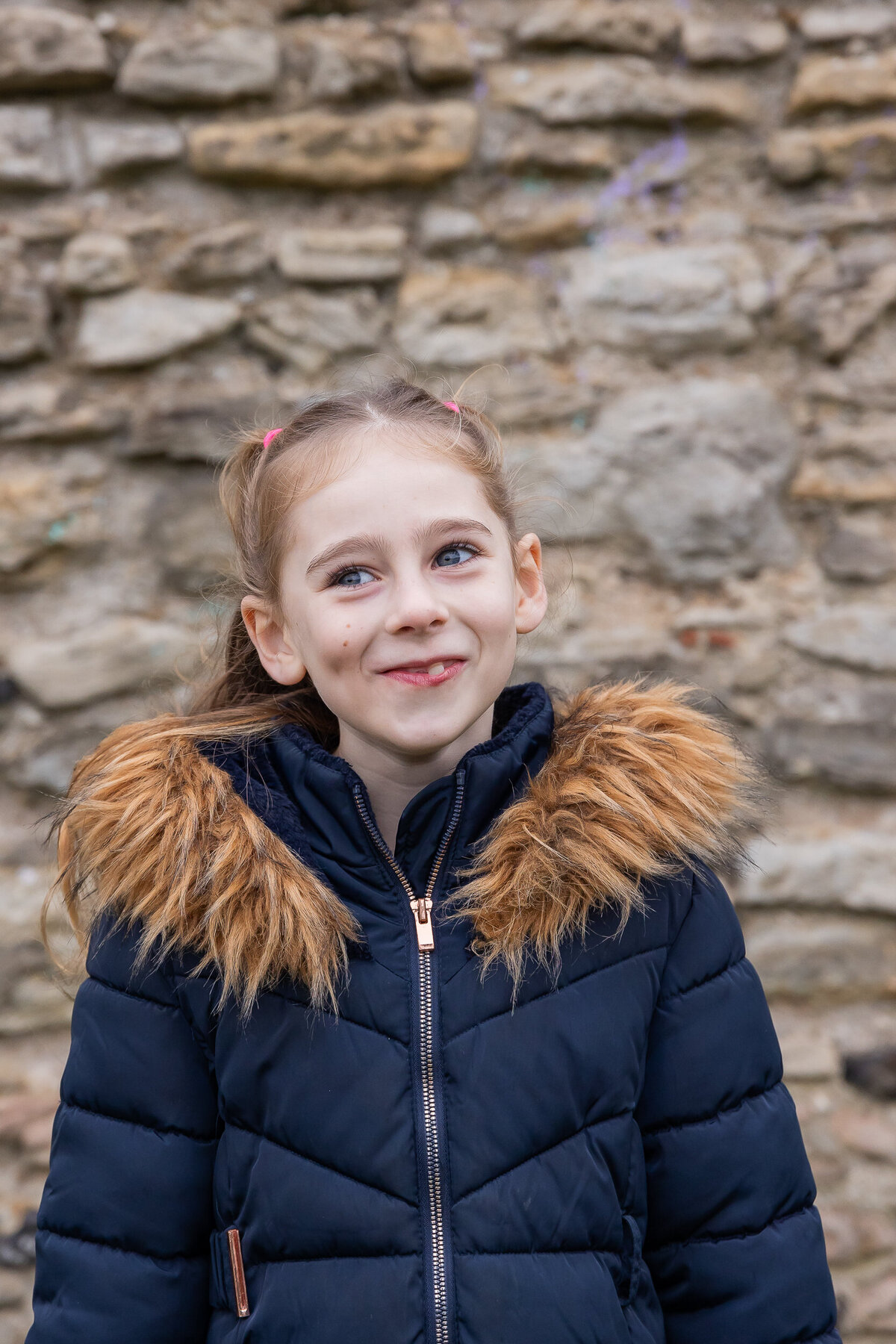 Young girl with a subtle smile, wearing a dark blue jacket with fur-lined hood, standing in front of a stone wall.