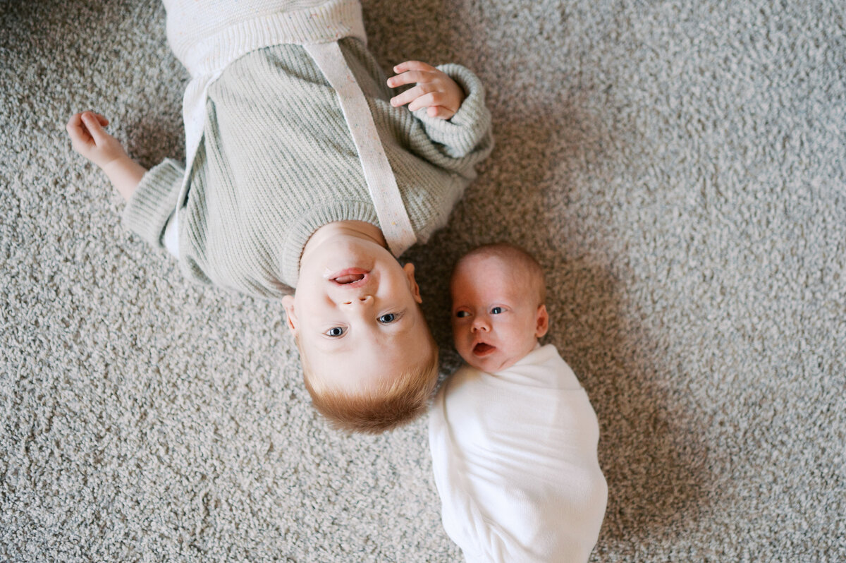 baby and big brother lie on the floor looking up at camera during newborn photos in benbrook fort worth