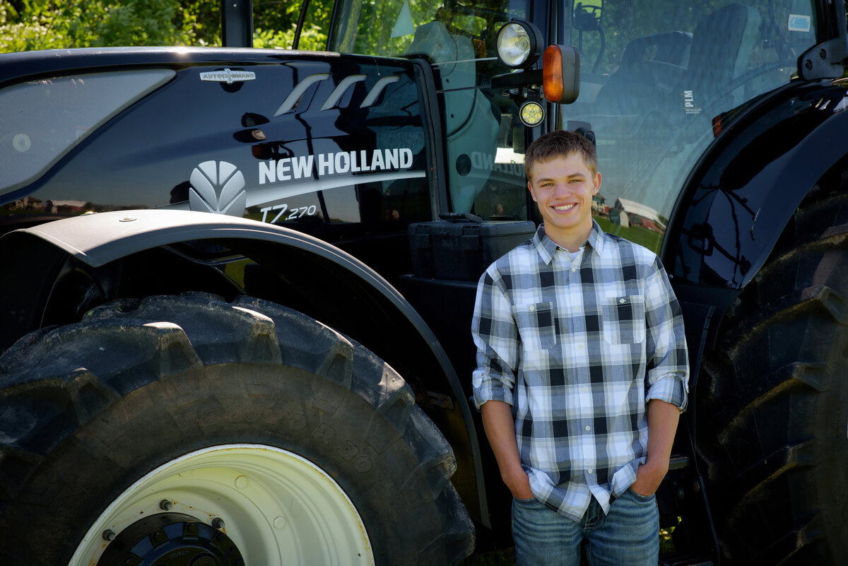 Luxemburg Casco high school senior boy wearing a black and white plaid button down shirt and jeans by a black New Holland tractor in farm field at his home near Green Bay, Wisconsin.