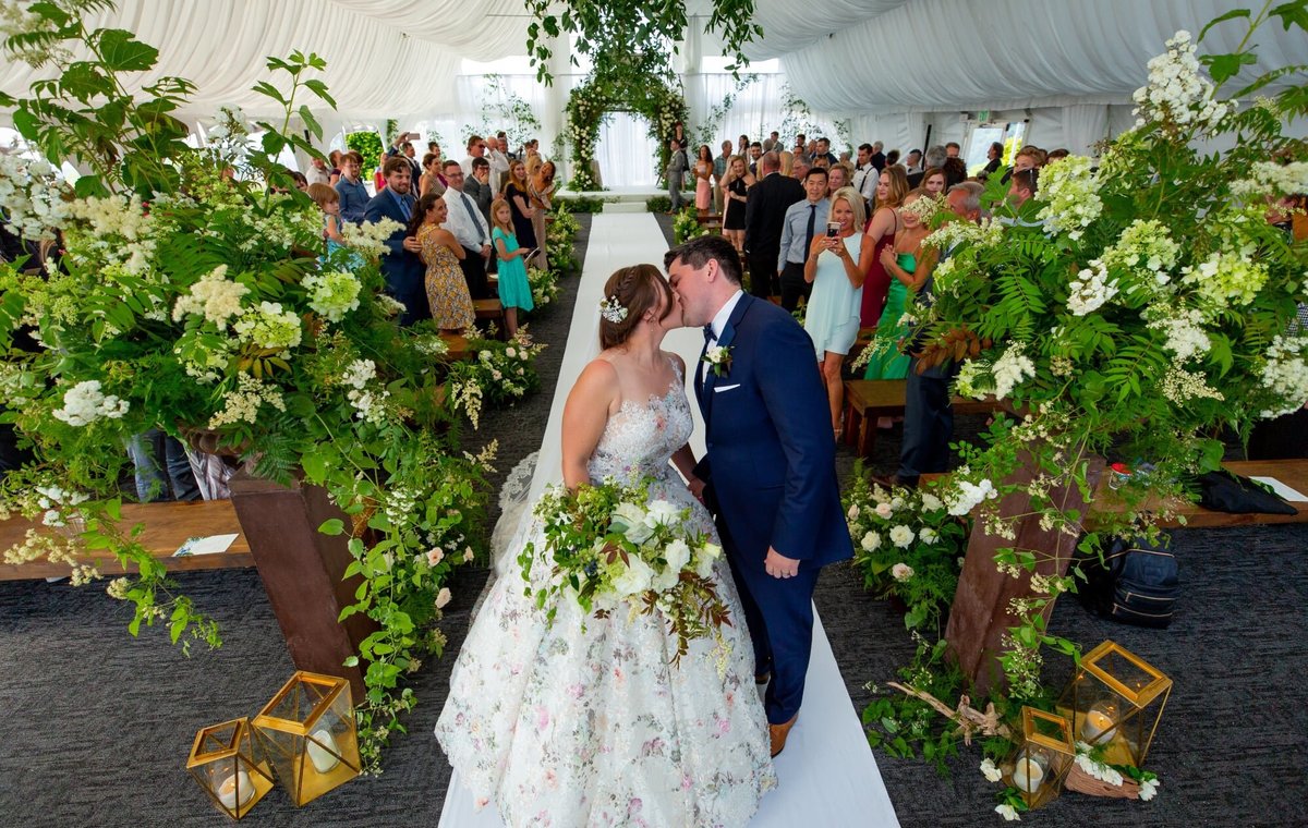 Bride and groom kissing on wedding ceremony aisle decorated with large h=greenery floral arrangements