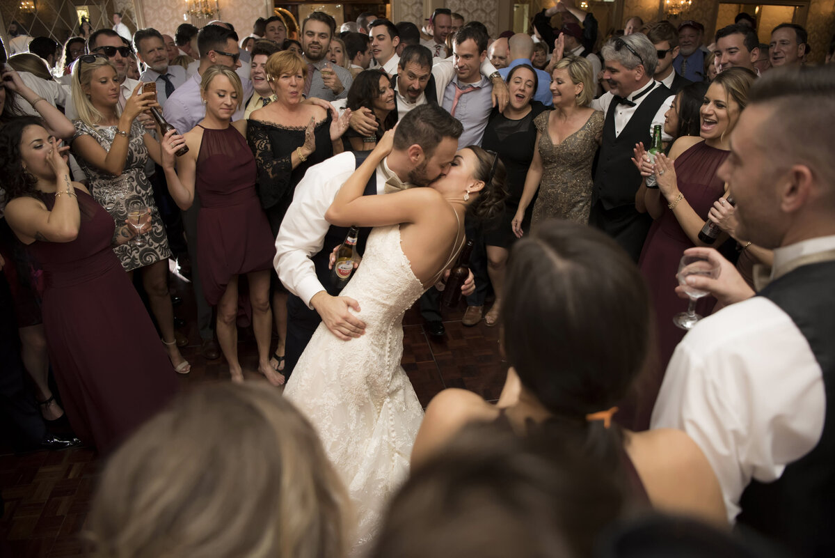 A bride and groom are surrounded by their wedding guests as they kiss at the end of their wedding day reception. 
