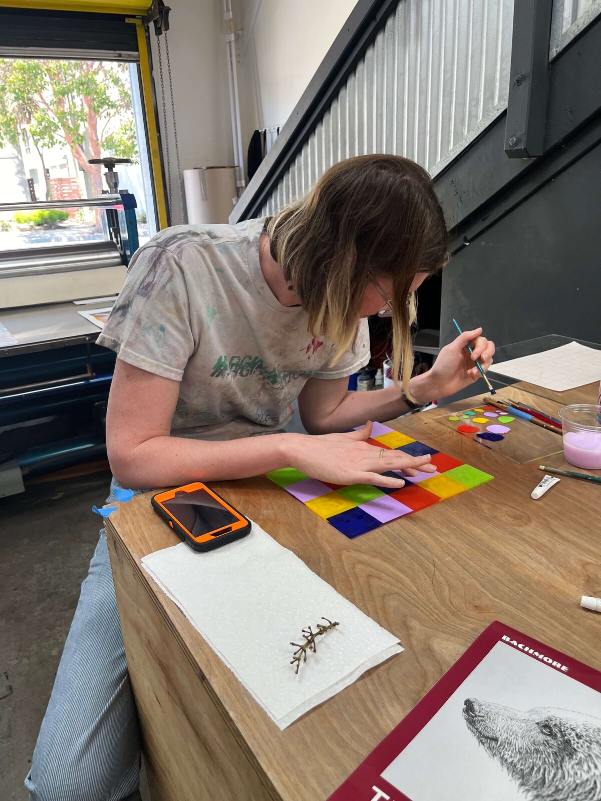 A participant sits at a studio table carefully painting a colorful grid on a plexiglass matrix as part of the monoprinting workshop.