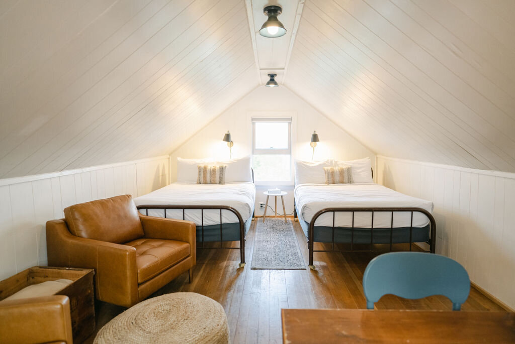 two beds frame the window in the attic floor of the Seagull Bay Cottage.  A brown leather armchair sits at the end of the bed on the left and a desk with a blue chair sits in the foreground.