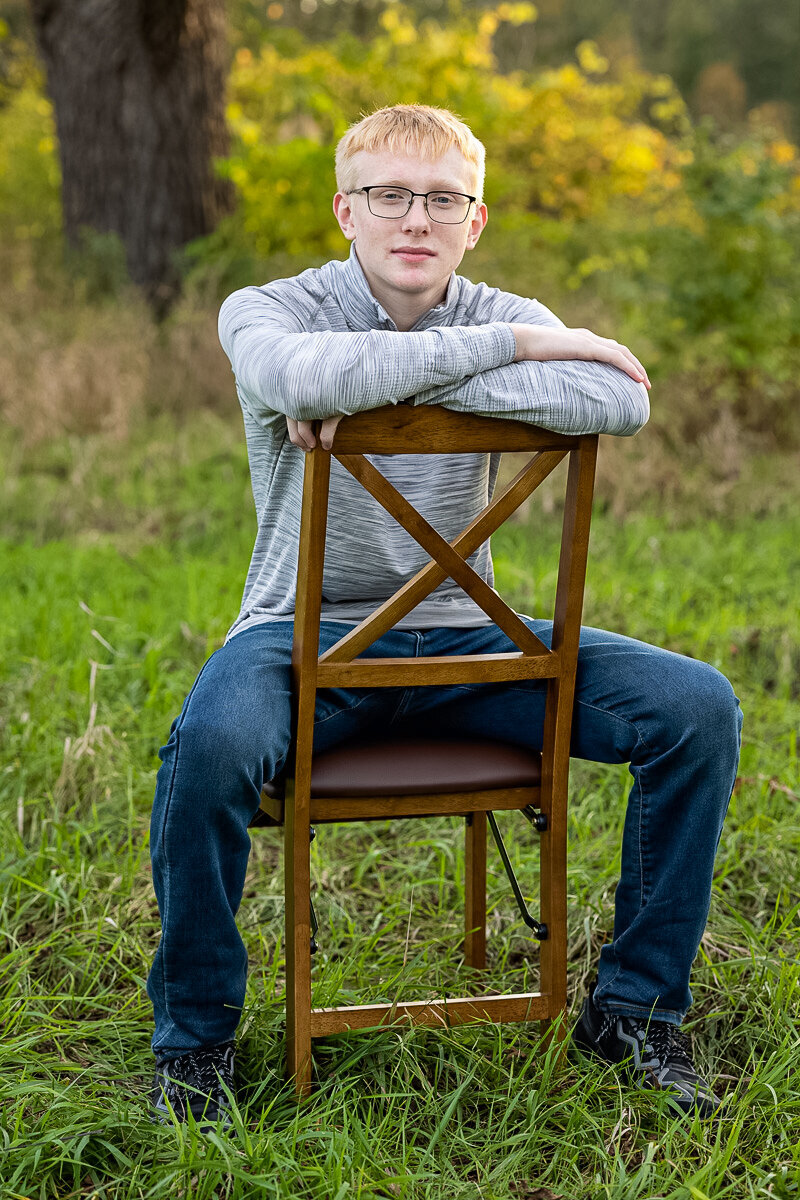 A male high school senior is posing seated backwards on a wooden chair.  The chair is in a field with a large tree in the distance.  He is wearing a gray shirt and jeans