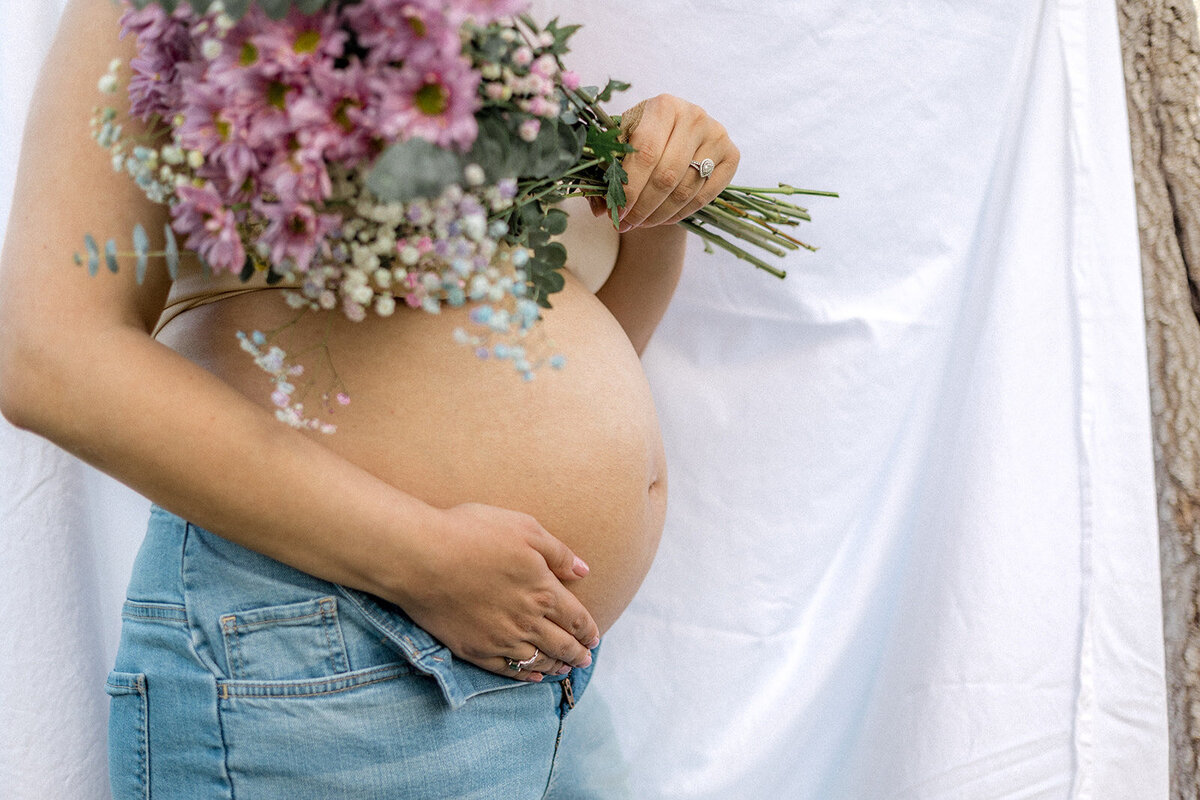A pregnant woman stands in front of a sheet outdoors while holding flowers