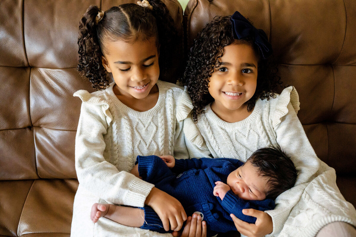 Twin girls holding newborn brother on couch