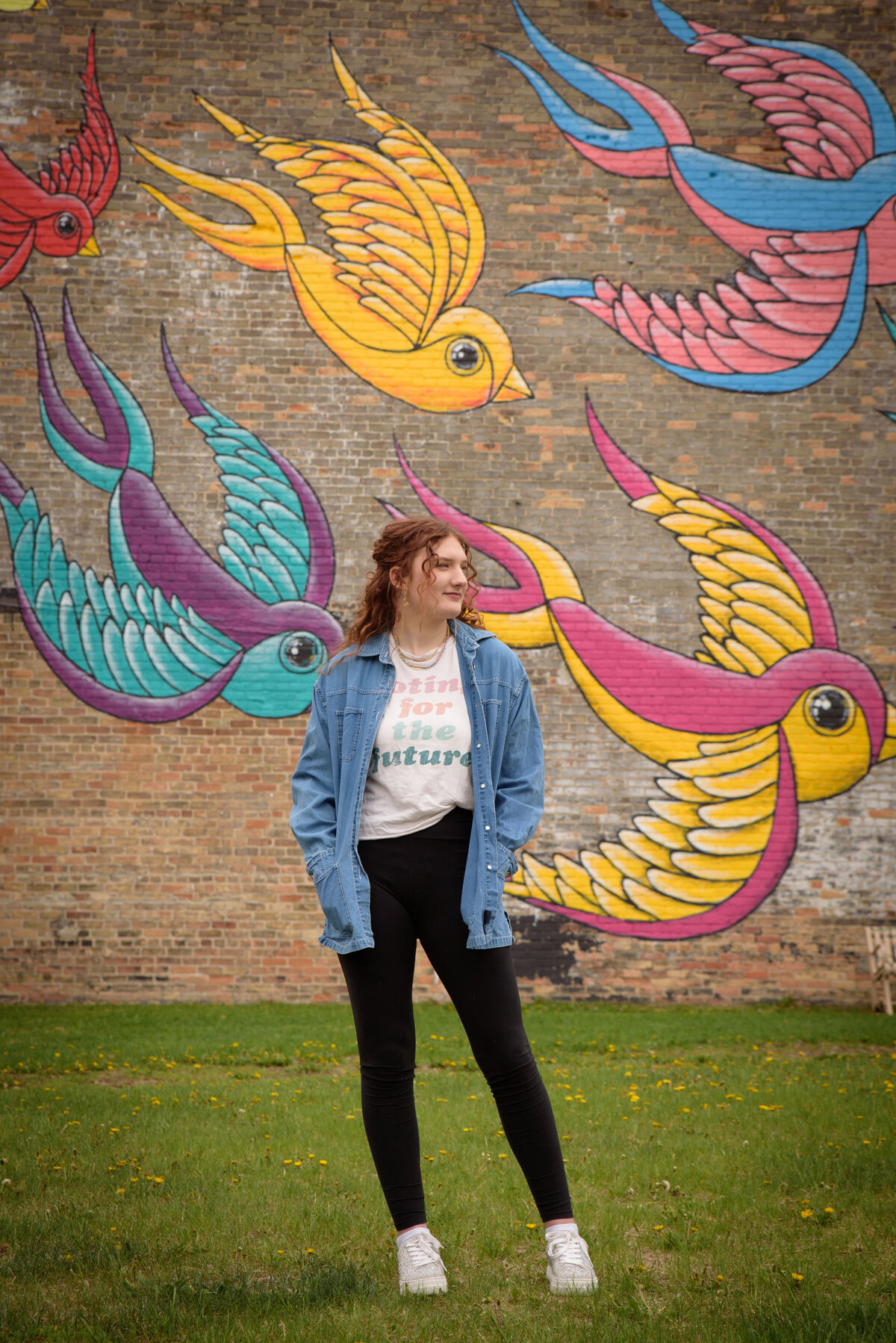 Green Bay East High School senior girl wearing a graphic t-shirt and leggings standing in front of a painted bird mural on a brick wall in an urban setting in downtown Green Bay, Wisconsin