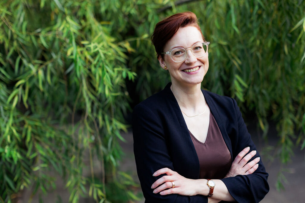 A person smiles with their arms crossed in front of greenery.